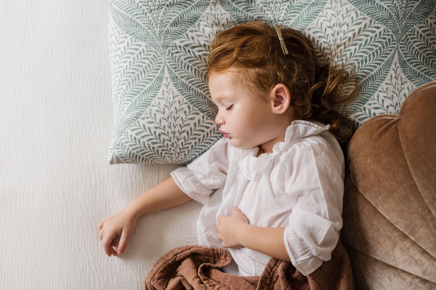 A small child in white ruffled clothing rests peacefully on a dusty pink velvet cushion.