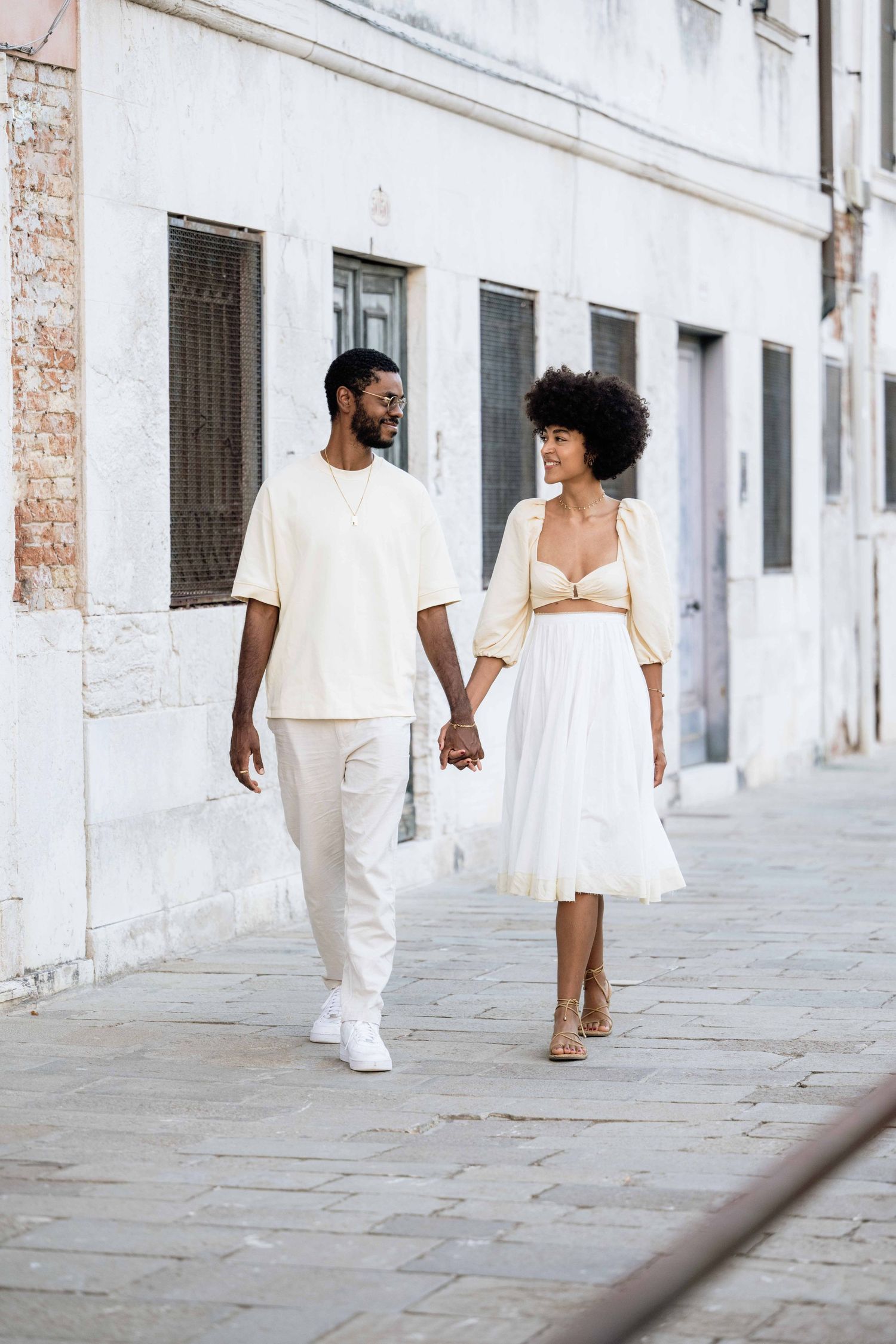 A couple in matching white outfits walking hand in hand along a historic street.