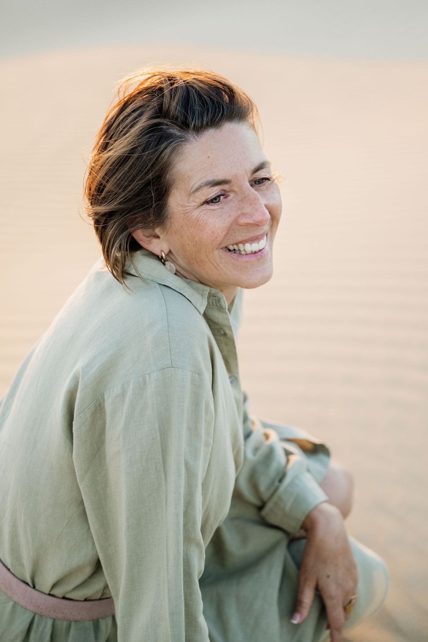 Woman in light green shirt smiling while posing on sandy dunes during golden hour.