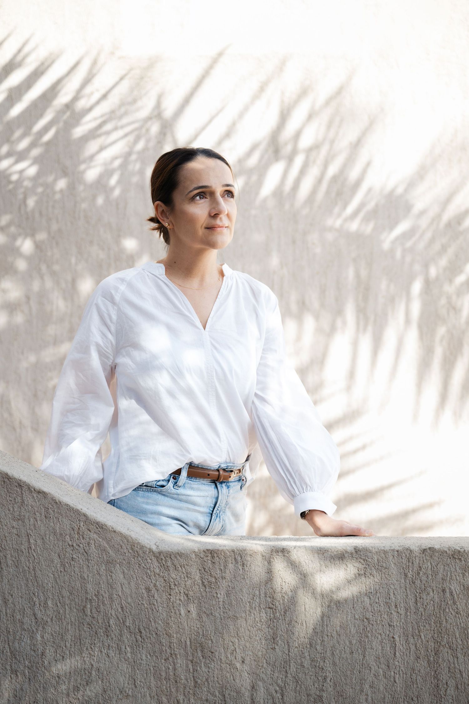 Woman in white button-down shirt and light jeans sits on concrete wall with palm tree shadows in background.