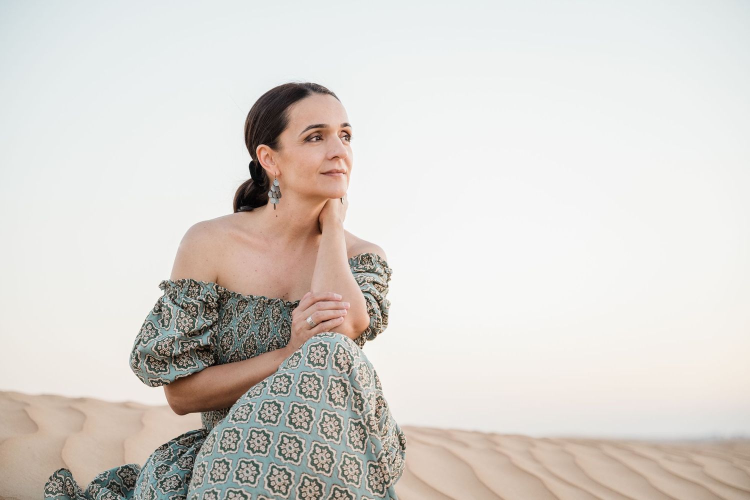 Woman in patterned dress poses against neutral background in natural light.
