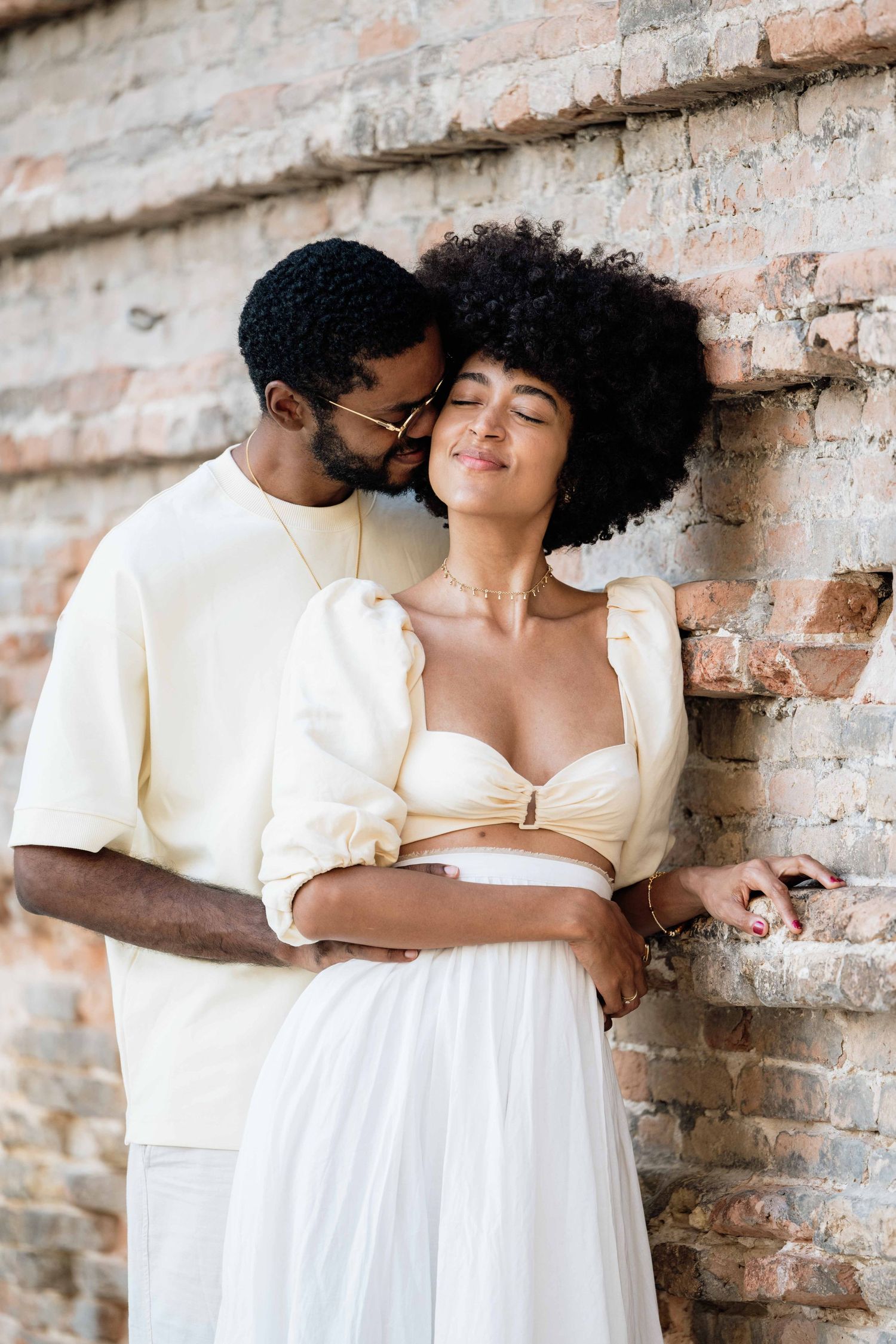 Romantic couple in cream and white outfits share intimate moments against brick wall.