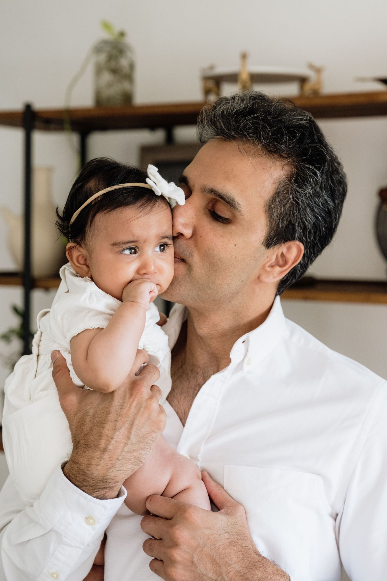 Intimate family moment with baby in white dress being held and kissed.