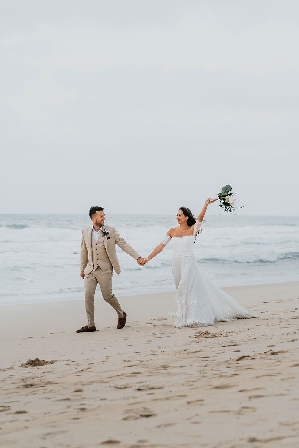 Playful newlyweds running and posing along a sandy beach with waves in the background.