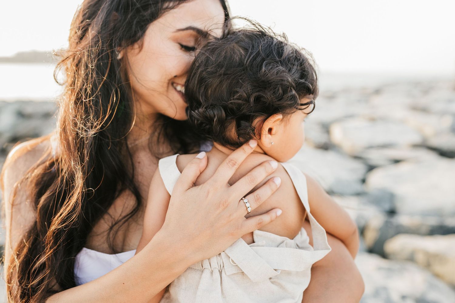 A tender family moment captured at sunset on a rocky beach with warm golden light and ocean waves in the background.