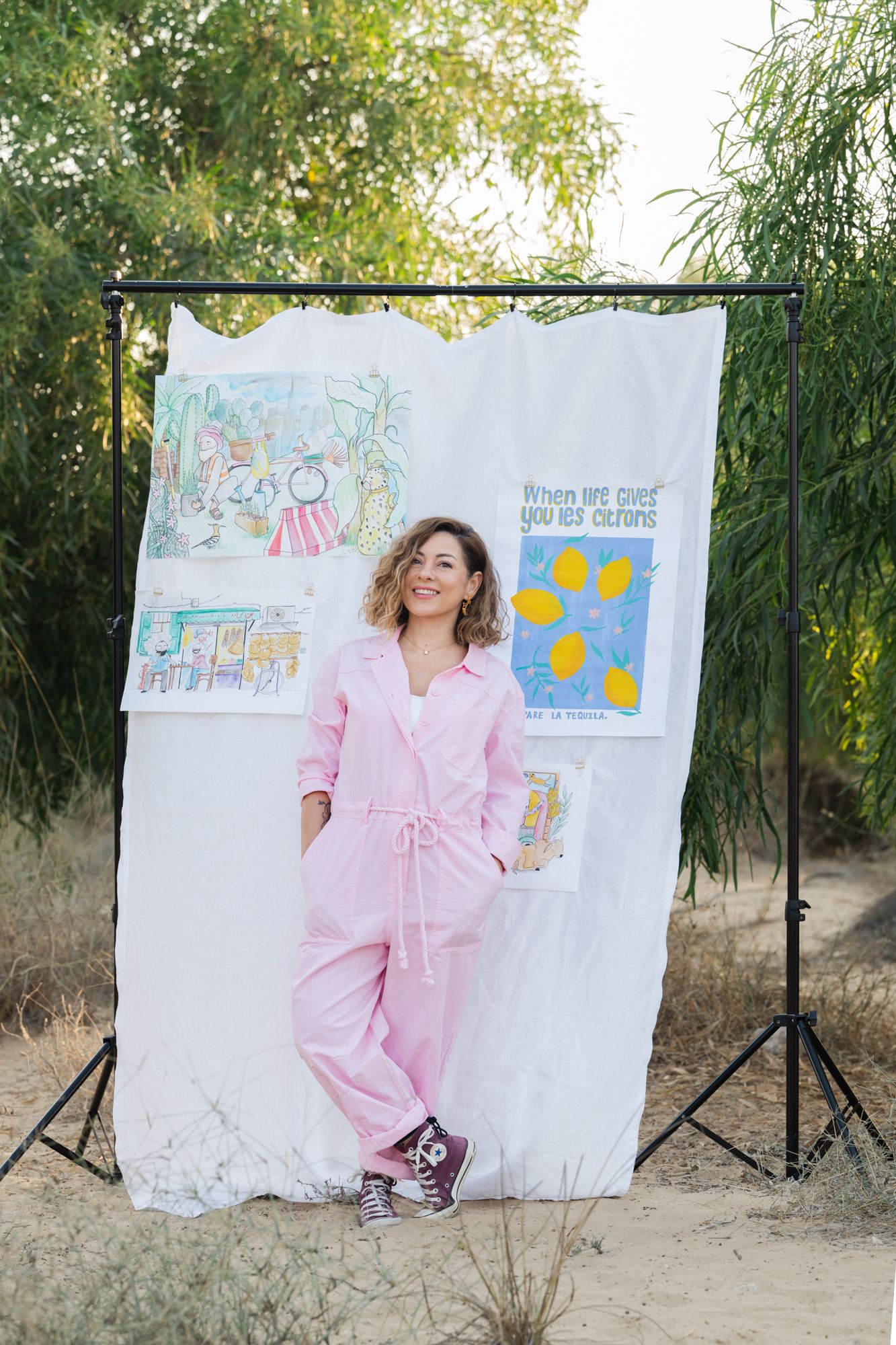 Woman wearing a pink jumpsuit poses playfully against a white backdrop with colorful artworks in an outdoor setting.