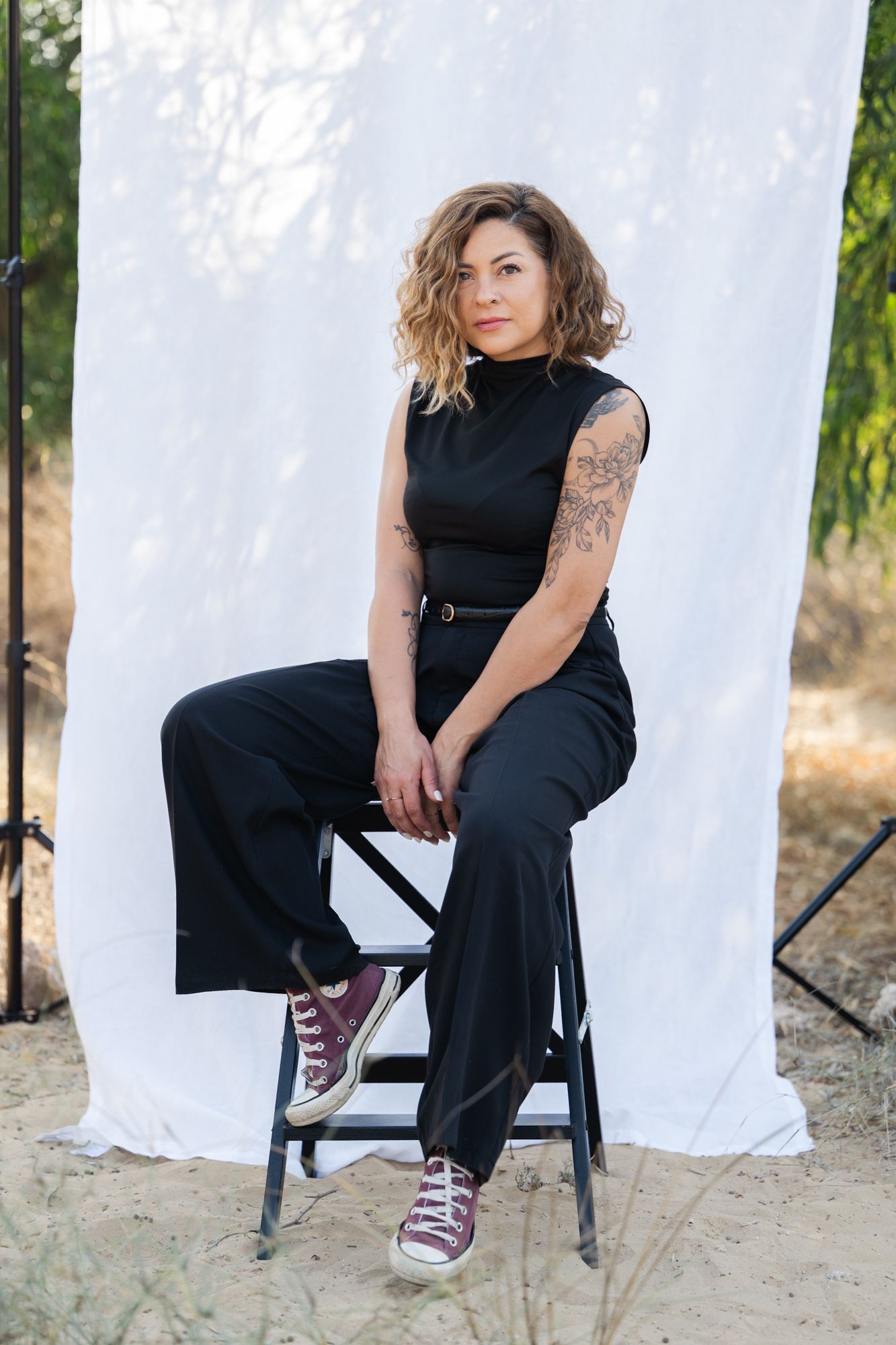 A person in a black outfit sits casually on a chair against a white backdrop wearing purple sneakers.