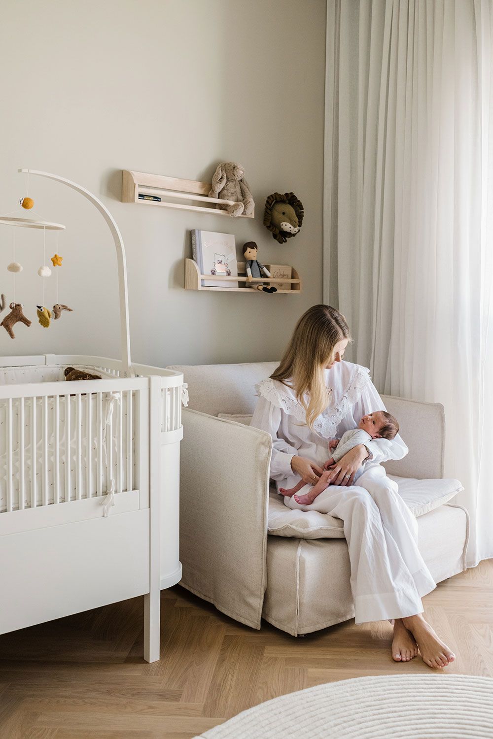 Mother and newborn baby in minimalist nursery room interior with white crib, armchair, and soft neutral decor in natural lighting.