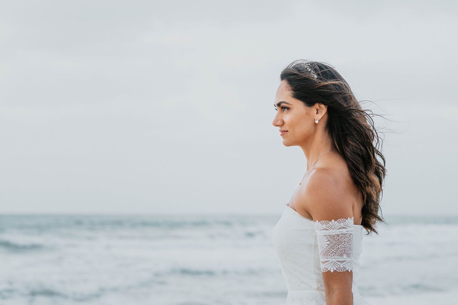 Bride in off-shoulder lace dress poses against serene beach backdrop.