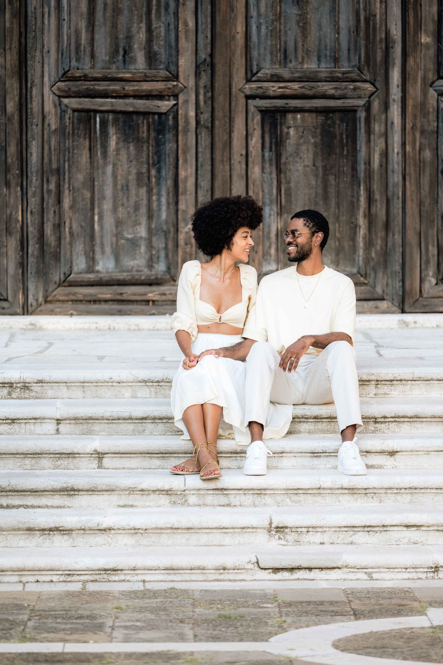 A couple shares a moment while sitting on stone steps in front of weathered wooden doors.