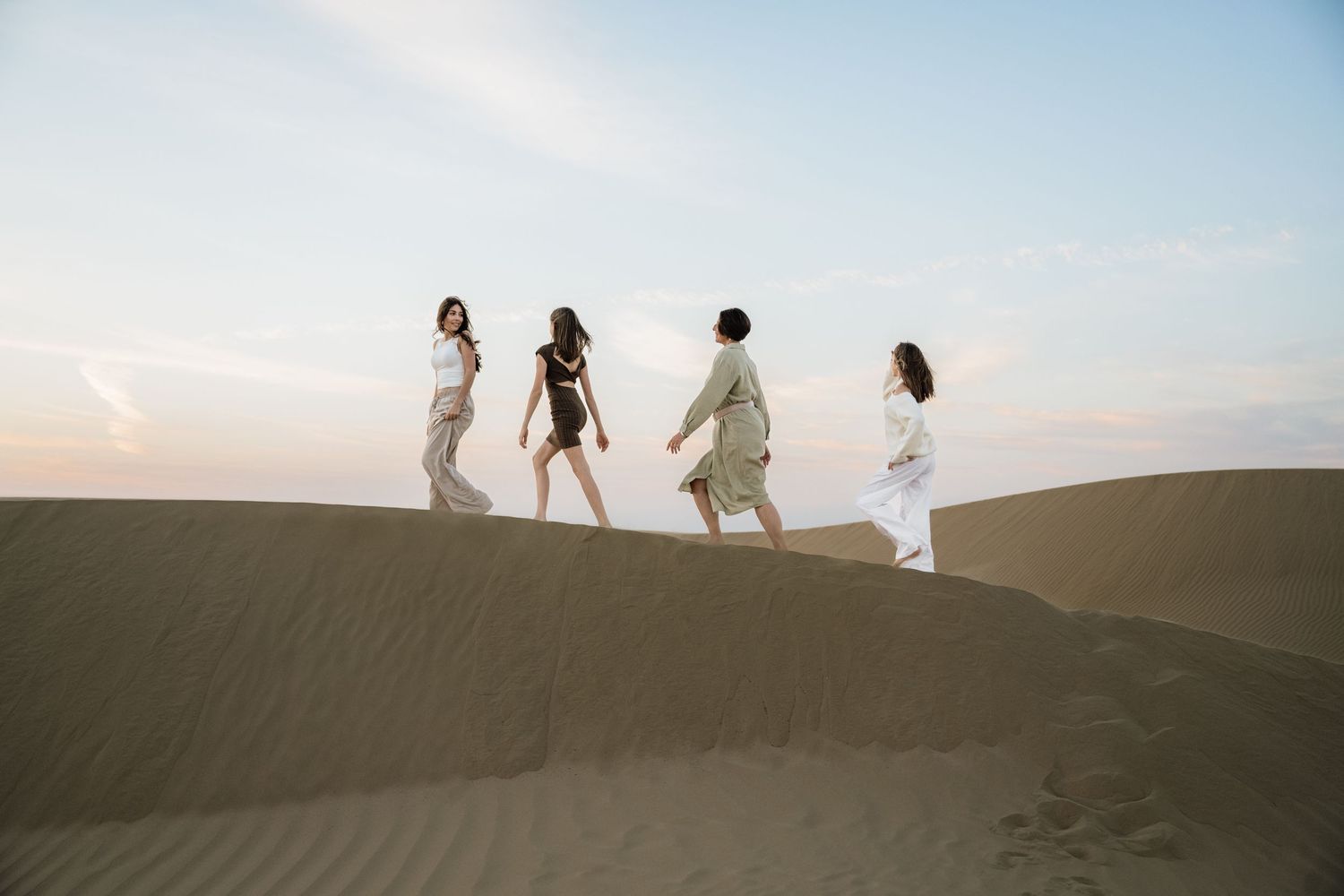 Silhouettes of people walking along the crest of sand dunes against a bright sky.
