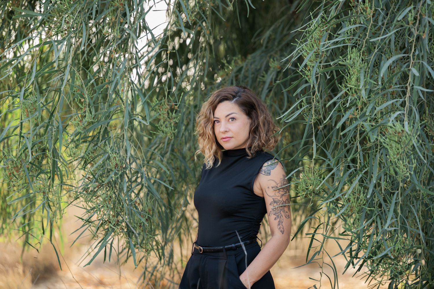 A woman in a black dress poses gracefully under drooping tree branches in a natural outdoor setting.