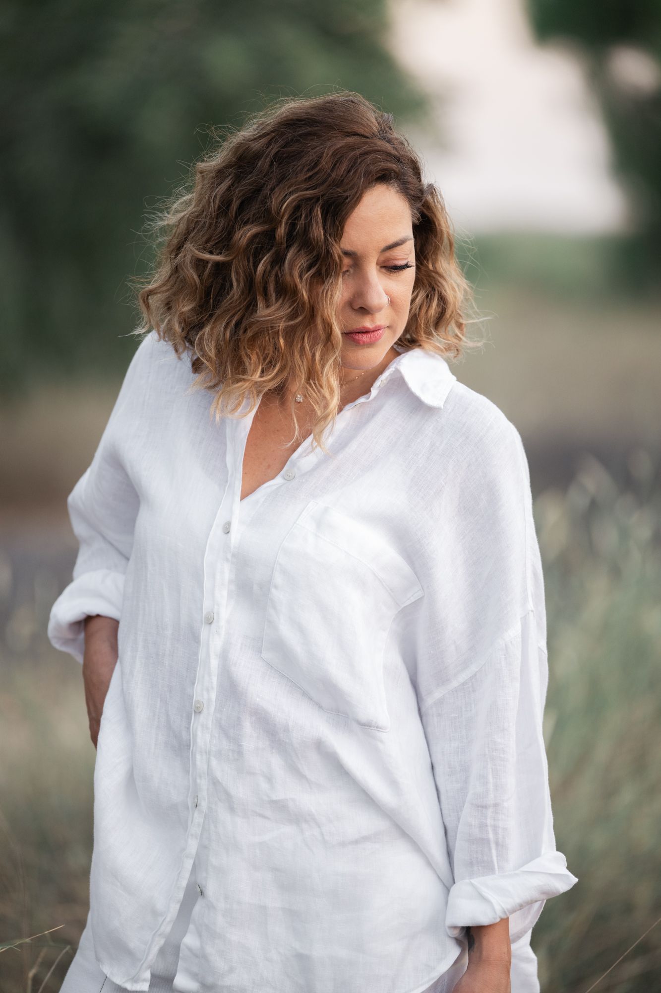 A woman in a white linen shirt poses outdoors in a natural setting with soft lighting and blurred background.