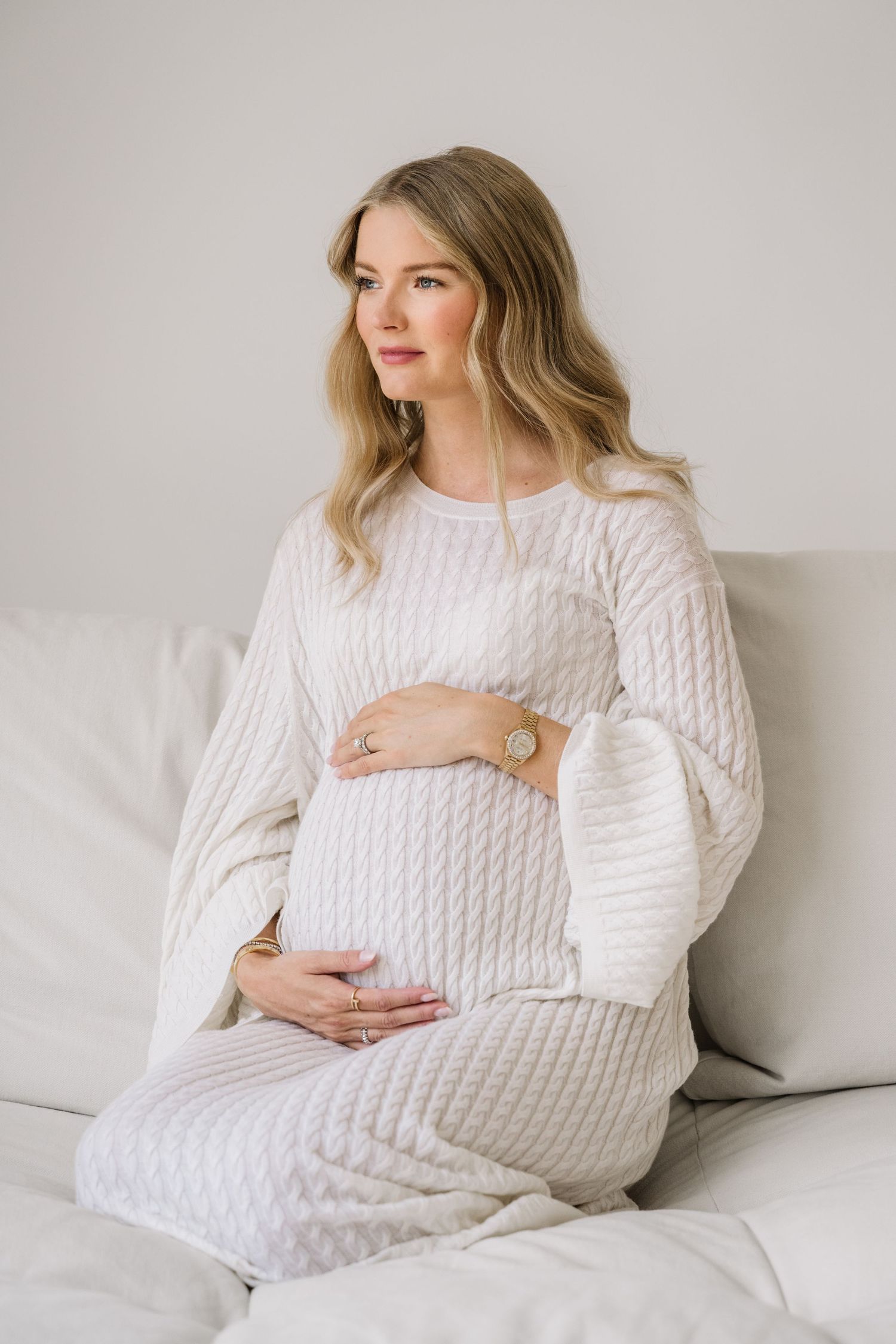 Maternity photos showing woman in white sweater on couch with growing belly.