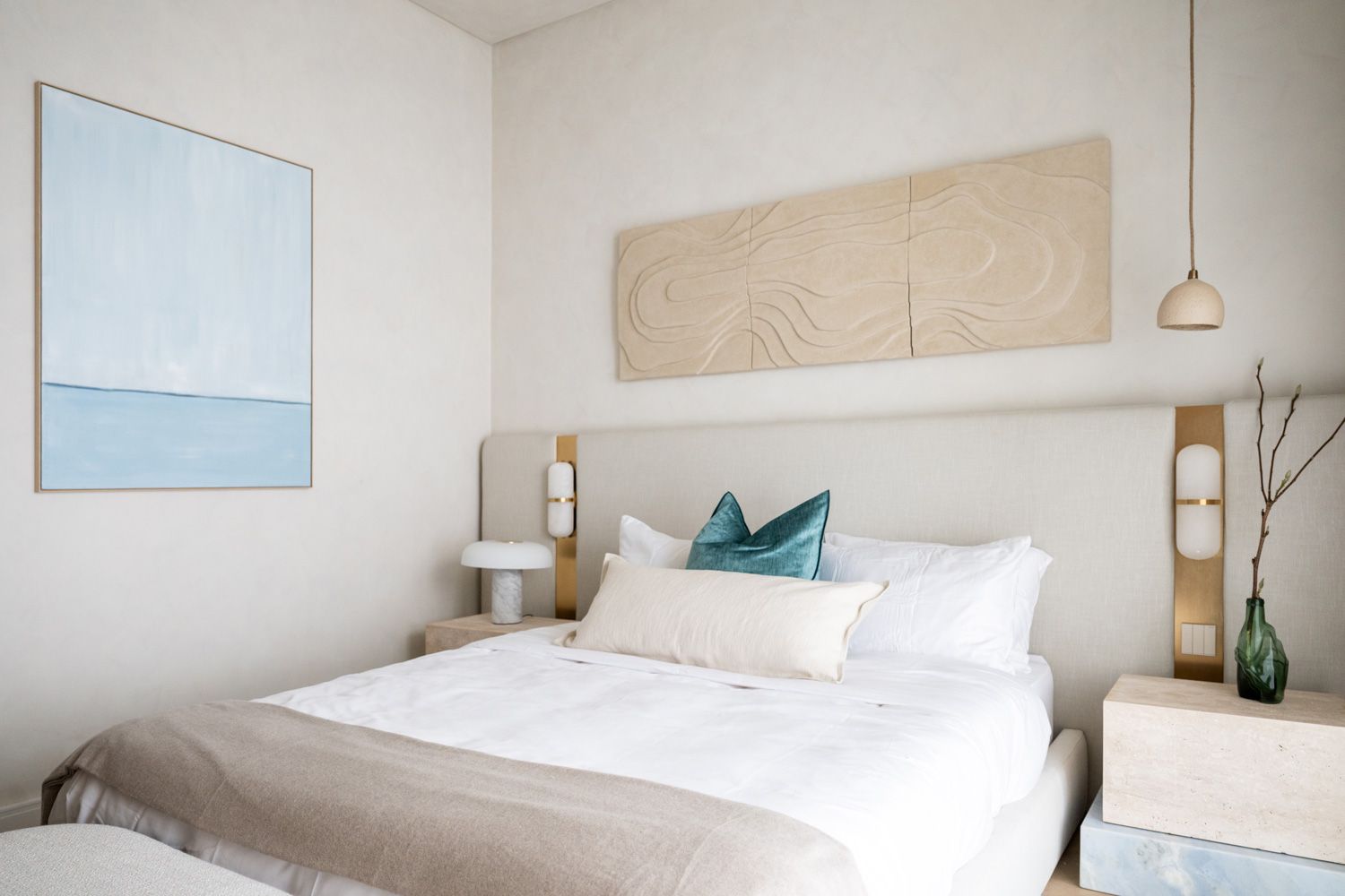 Modern minimalist bedroom with white bedding, teal accent pillow, neutral artwork, and wall-mounted sconces.