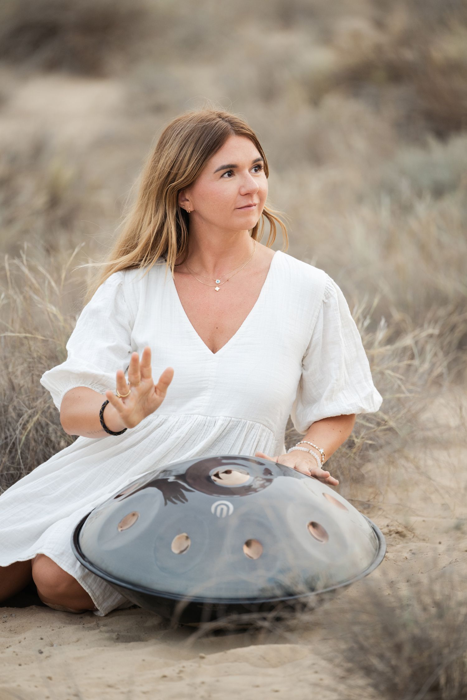 A person in a white dress sits on the ground with a handpan drum in a desert landscape at sunset.