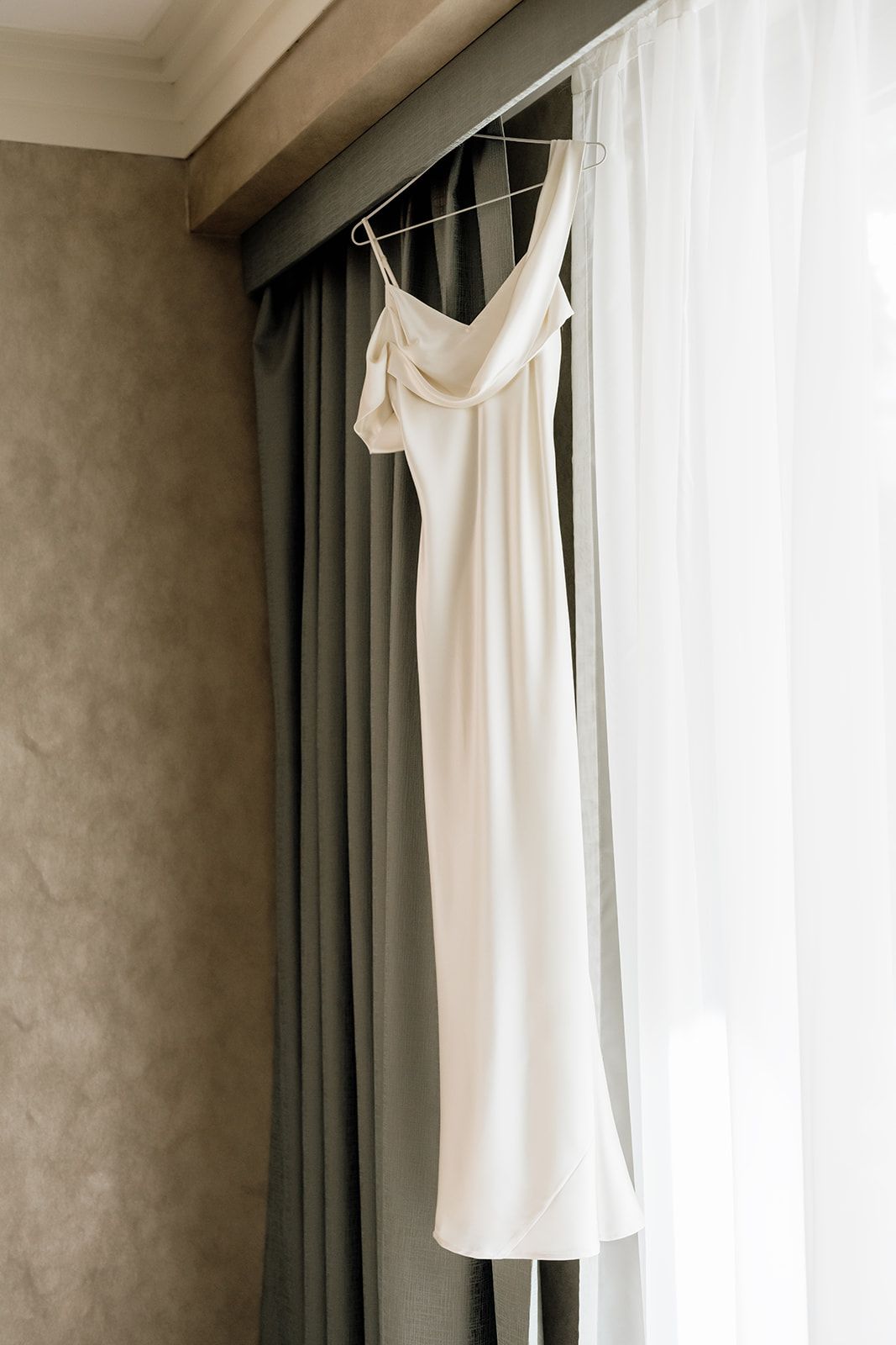 White wedding dress hanging by window with soft natural light and grey curtains.