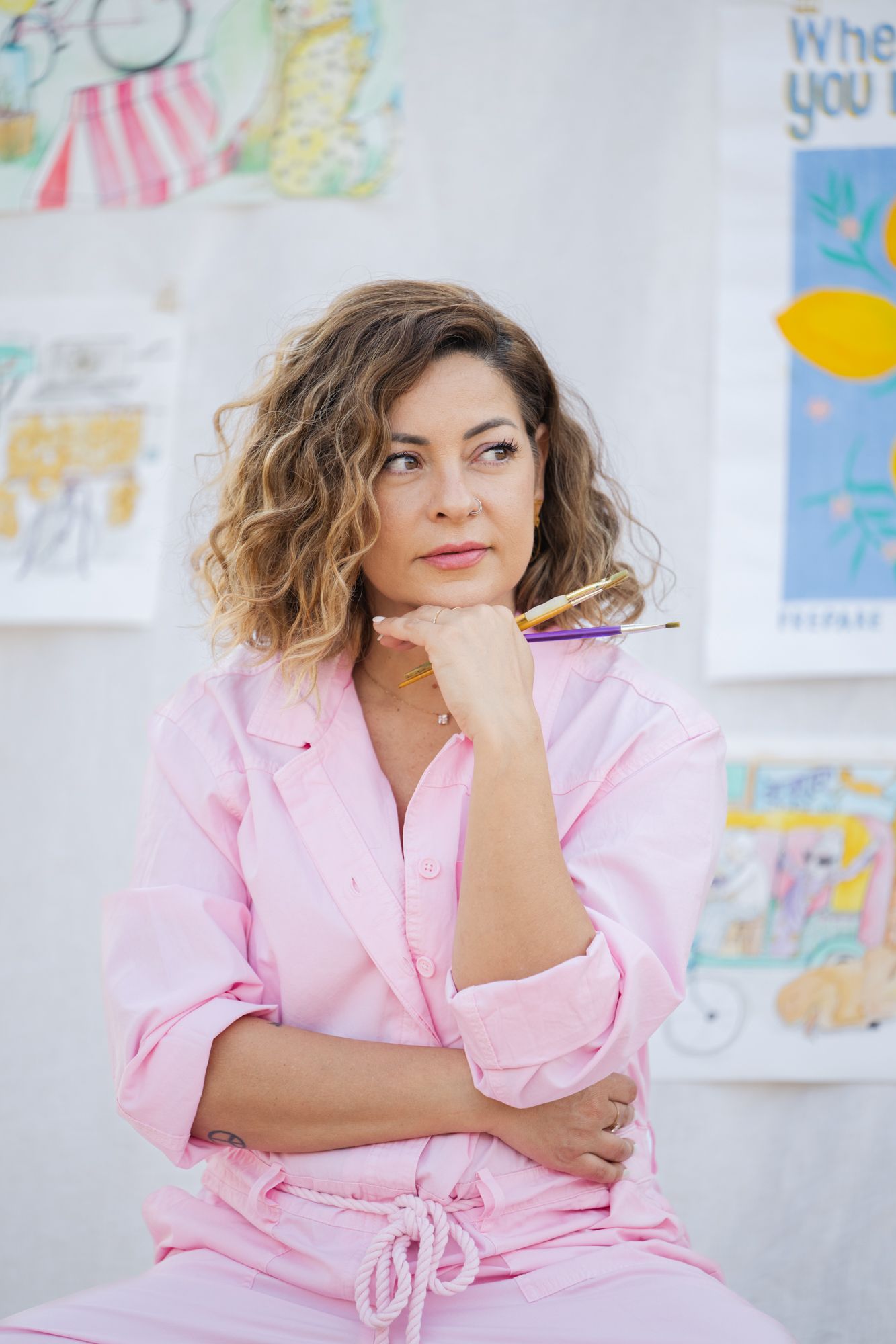 Woman wearing pink jumpsuit poses thoughtfully against white backdrop with artworks in background.