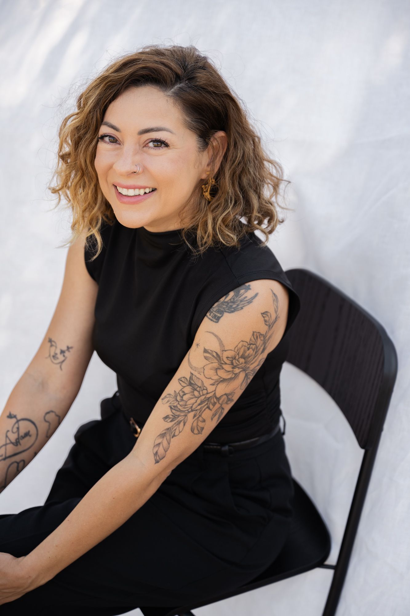 A lady in black attire sits on a chair with a relaxed demeanor, displaying artistic tattoos against a white background.