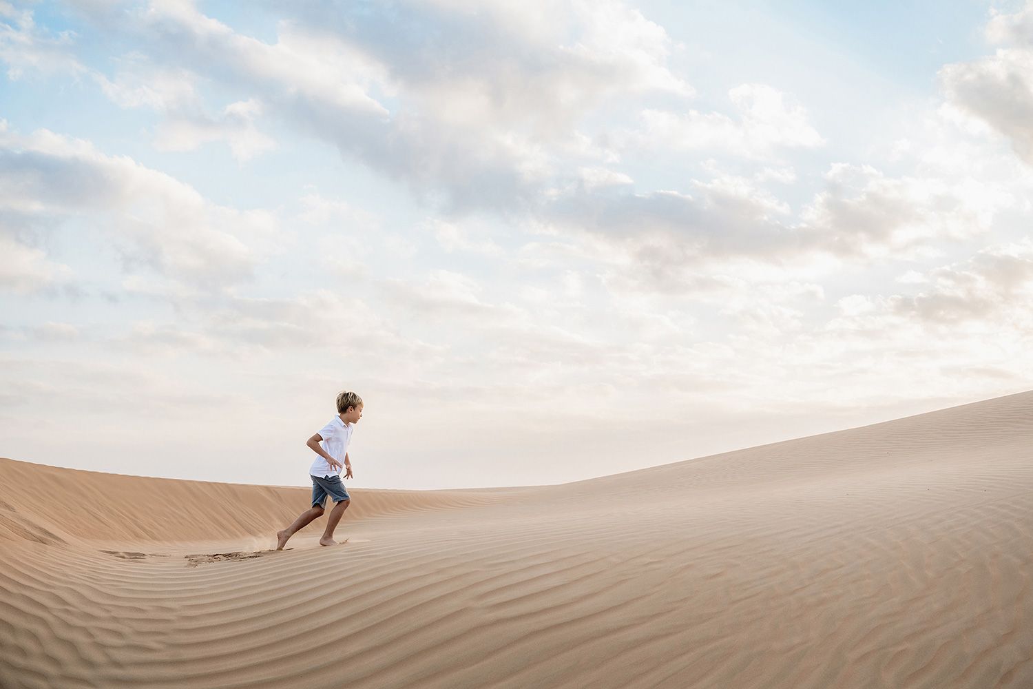 Person running across vast desert sand dunes at sunset with dramatic sky and rippling sand patterns.
