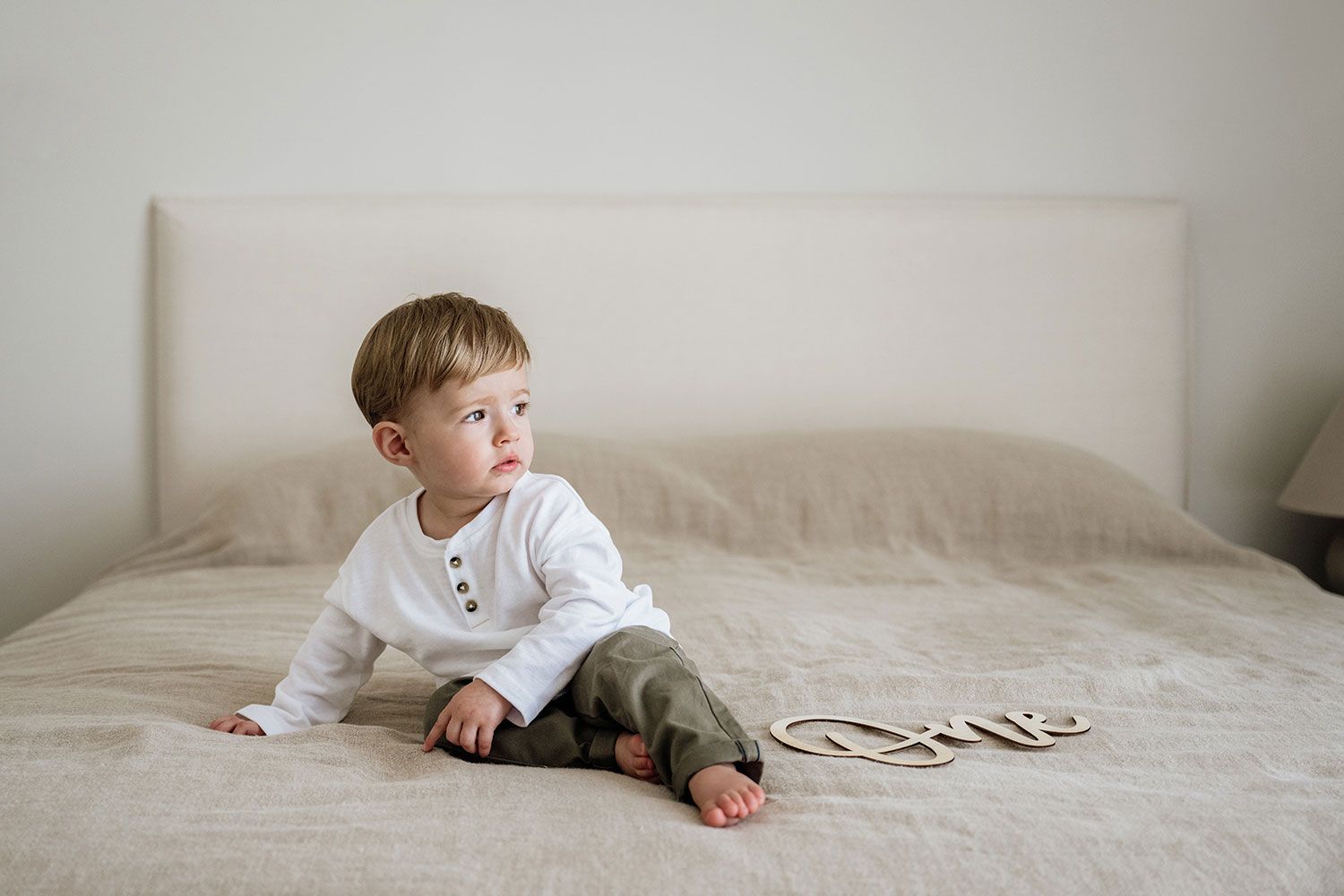 A baby in a white sweater crawls on a white bed while looking off to the side.