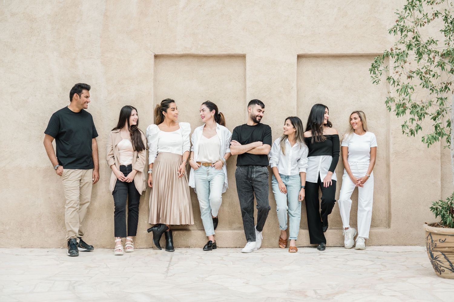 Group in casual outfits poses against a neutral beige wall in an outdoor setting.