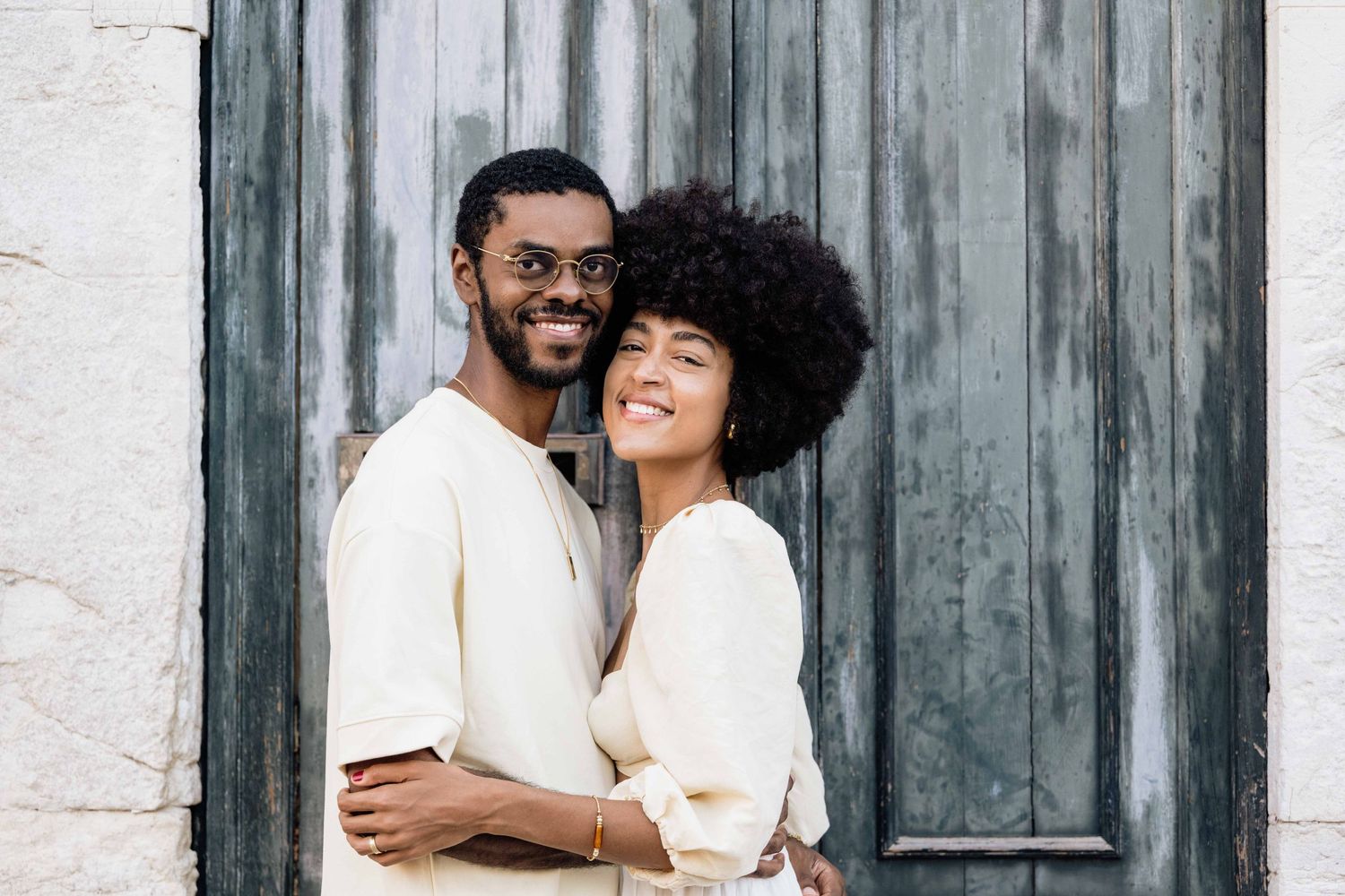 A romantic couple dressed in white outfits embrace against a rustic wall background.