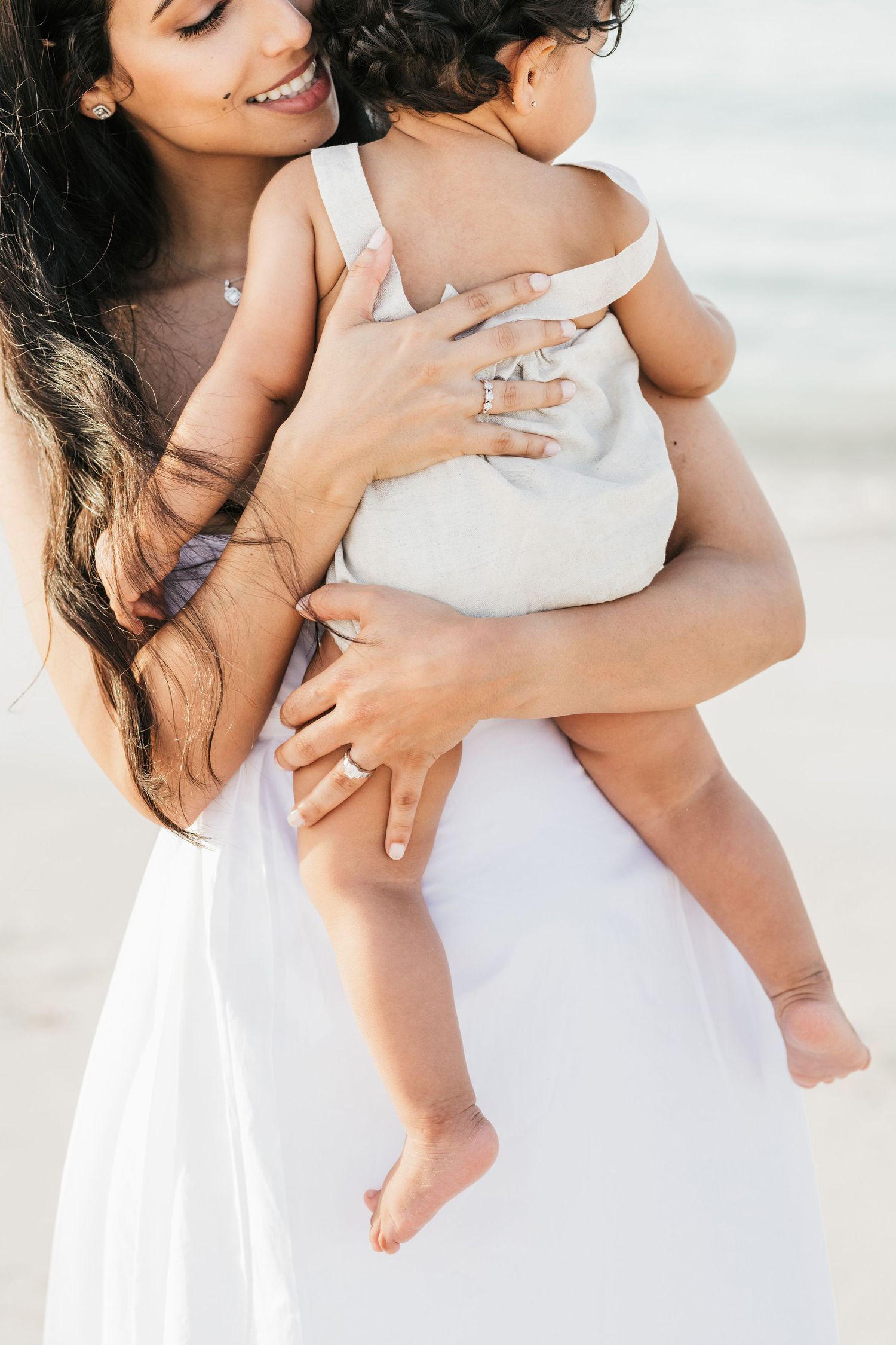 Parent in white dress holding and embracing small child against beach background in soft lighting.