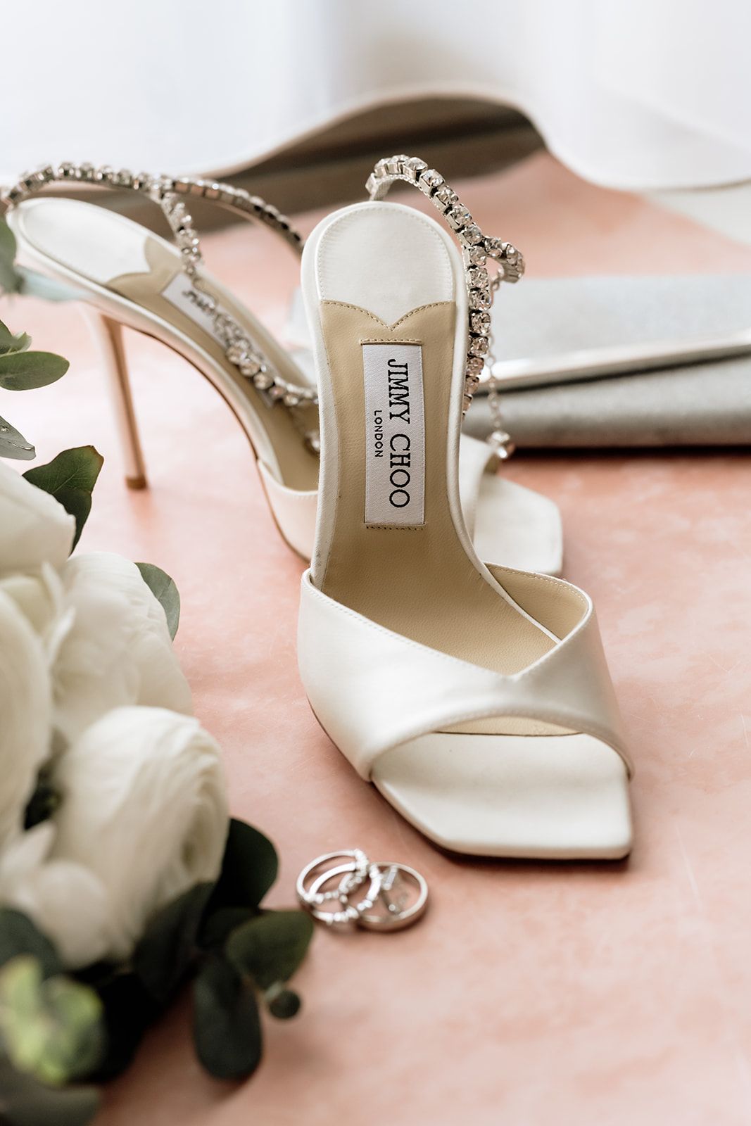 Elegant white bridal shoes with crystal details and roses on pink surface.