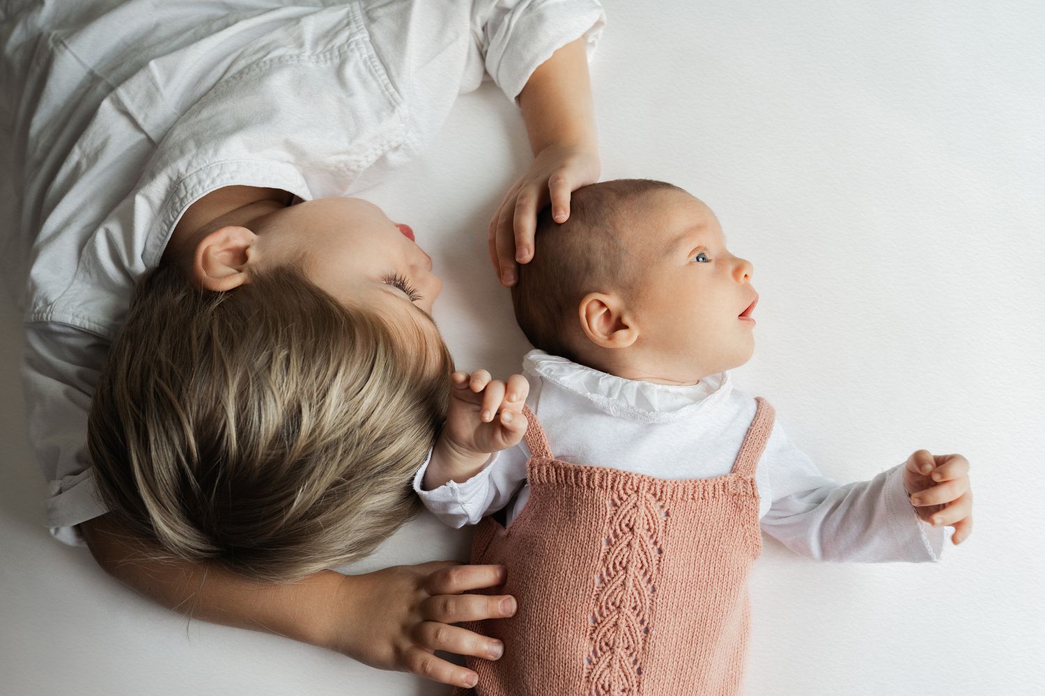 Mother lying beside her newborn baby in a white knit outfit, gently touching the infant's head in a tender moment.