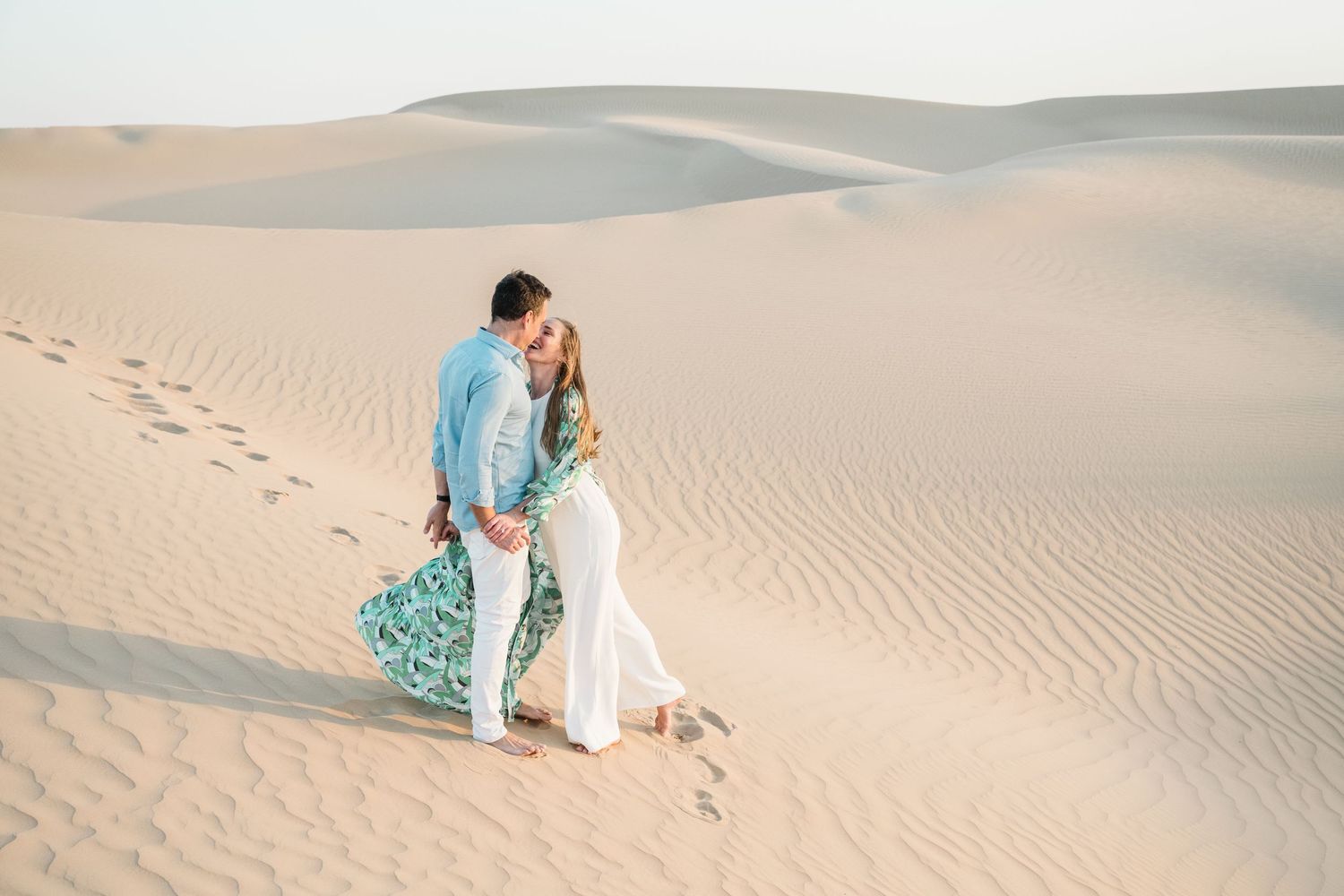Romantic desert photoshoot featuring couple sitting and walking on sand dunes.