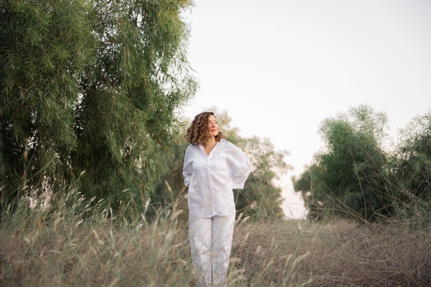 A series of seven connected outdoor photos showing a woman in white walking through tall grass and trees at sunset.