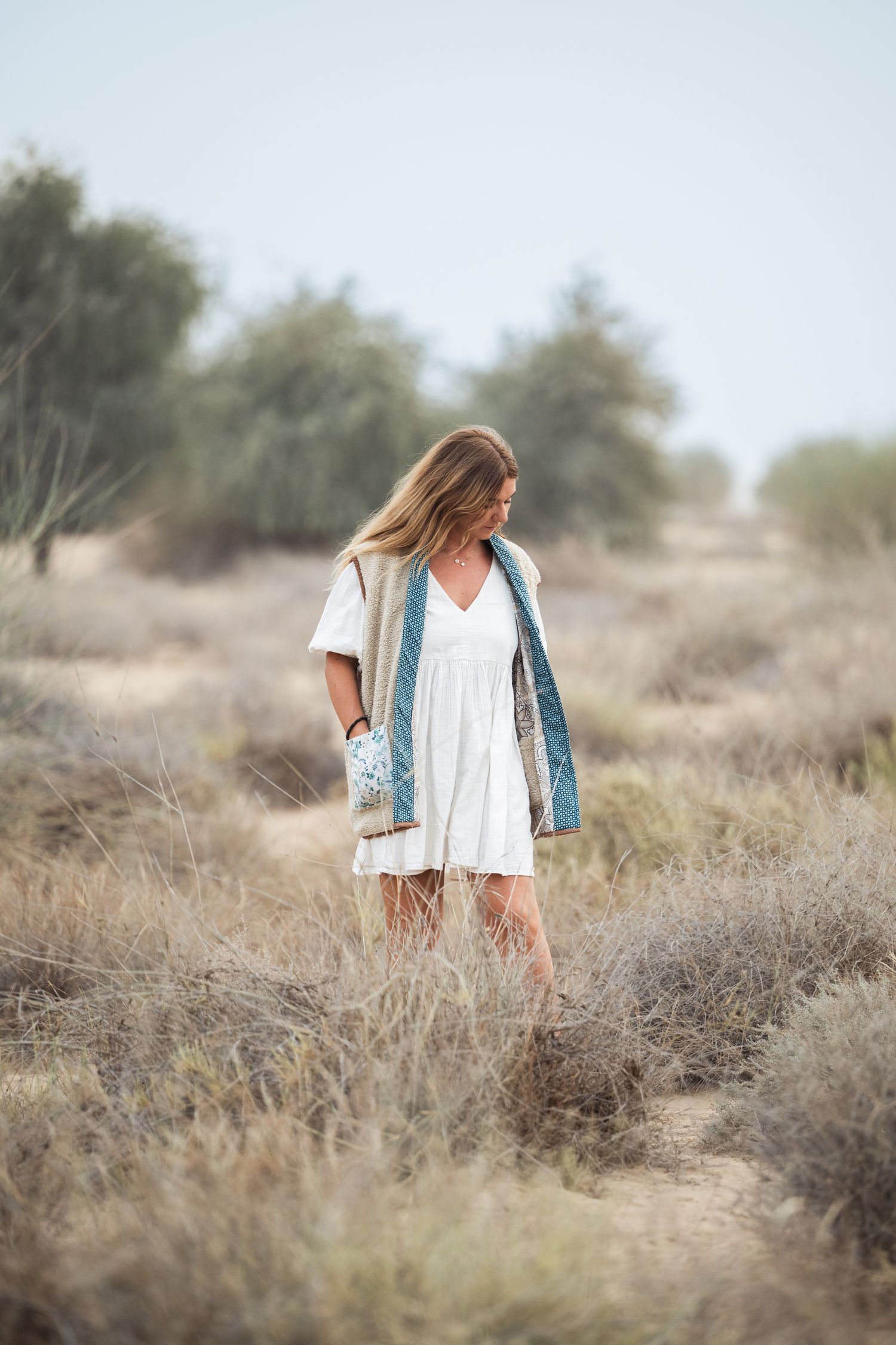 A person in a white dress and blue cardigan stands thoughtfully in a desert landscape with dry brush and soft lighting.