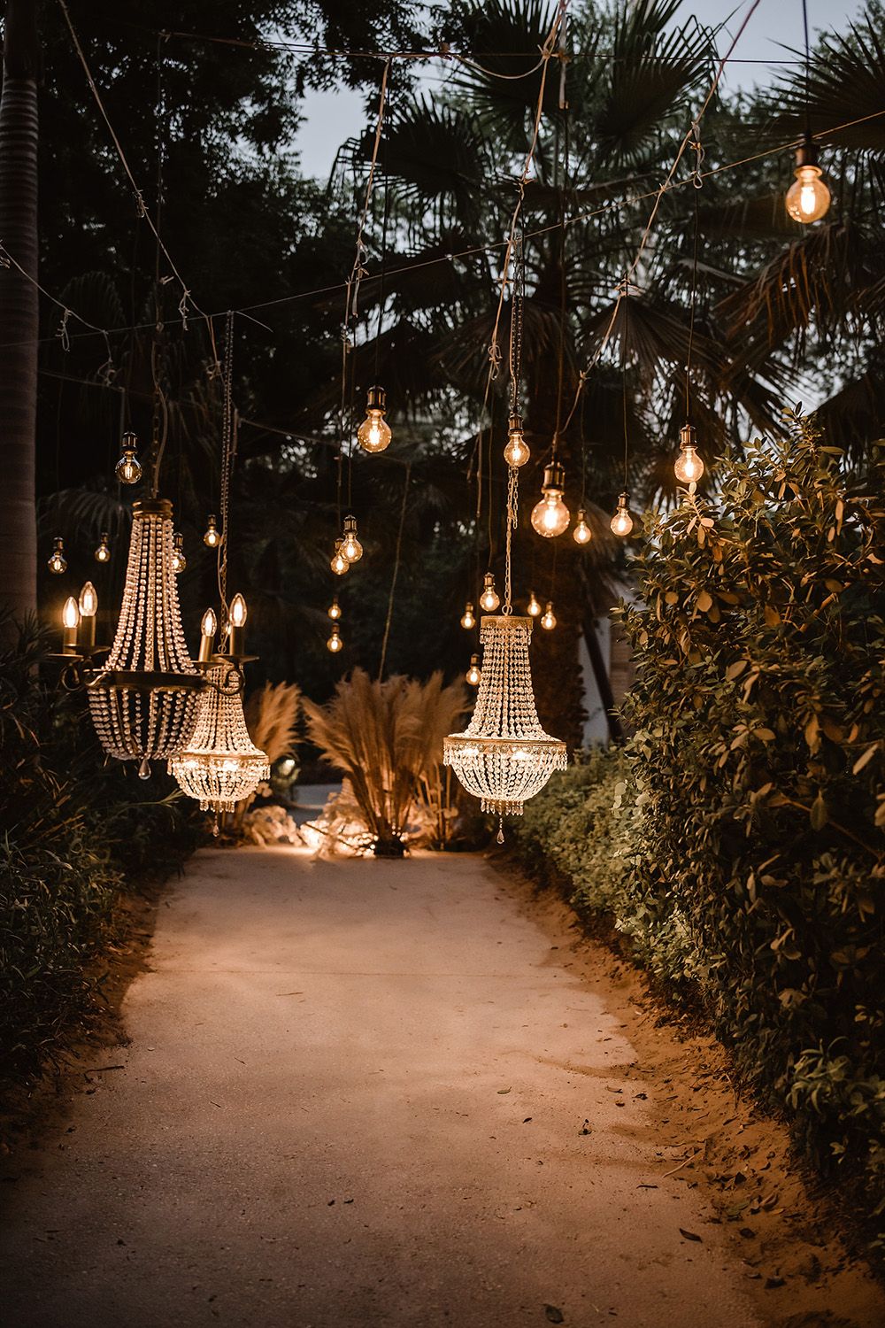 Illuminated garden path with crystal chandeliers and string lights creating magical ambiance.
