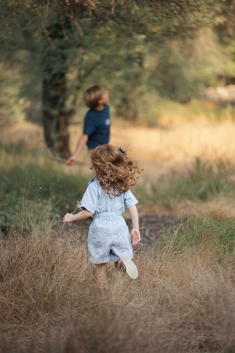 Child in light blue overalls runs through tall grass in a dreamy outdoor setting at golden hour.