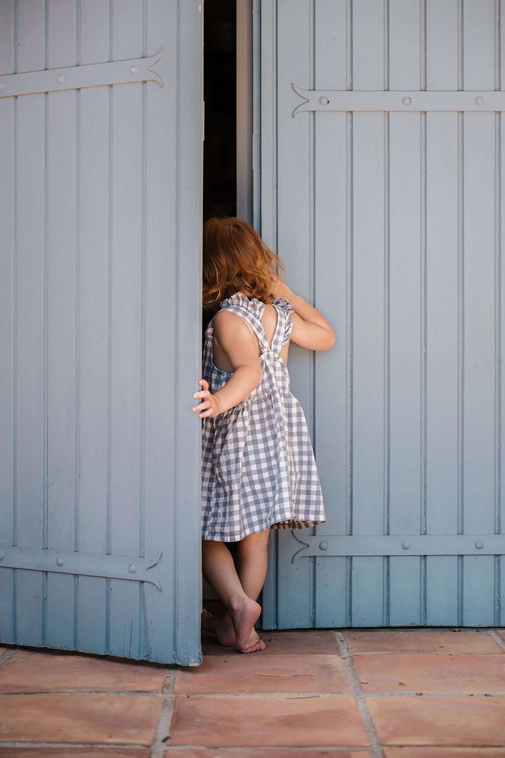 A child in a checkered dress peeks through a partially open blue door while playing hide and seek.