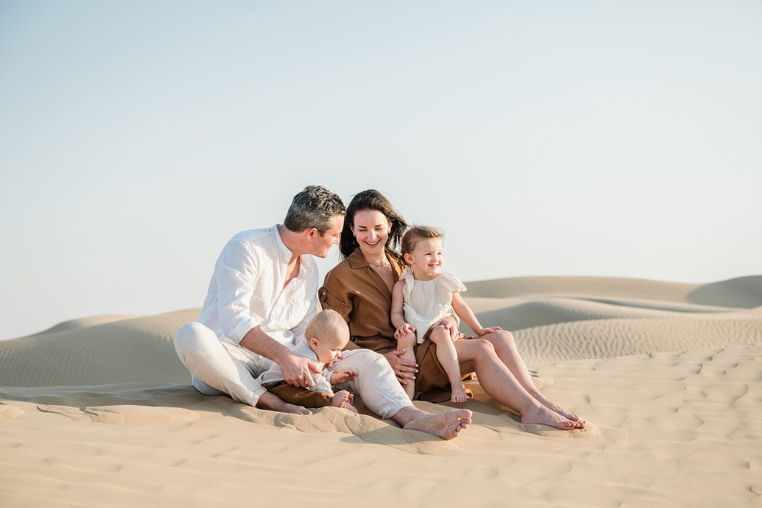 Family lounging together on sandy dunes in desert landscape at sunset wearing neutral colored clothing.