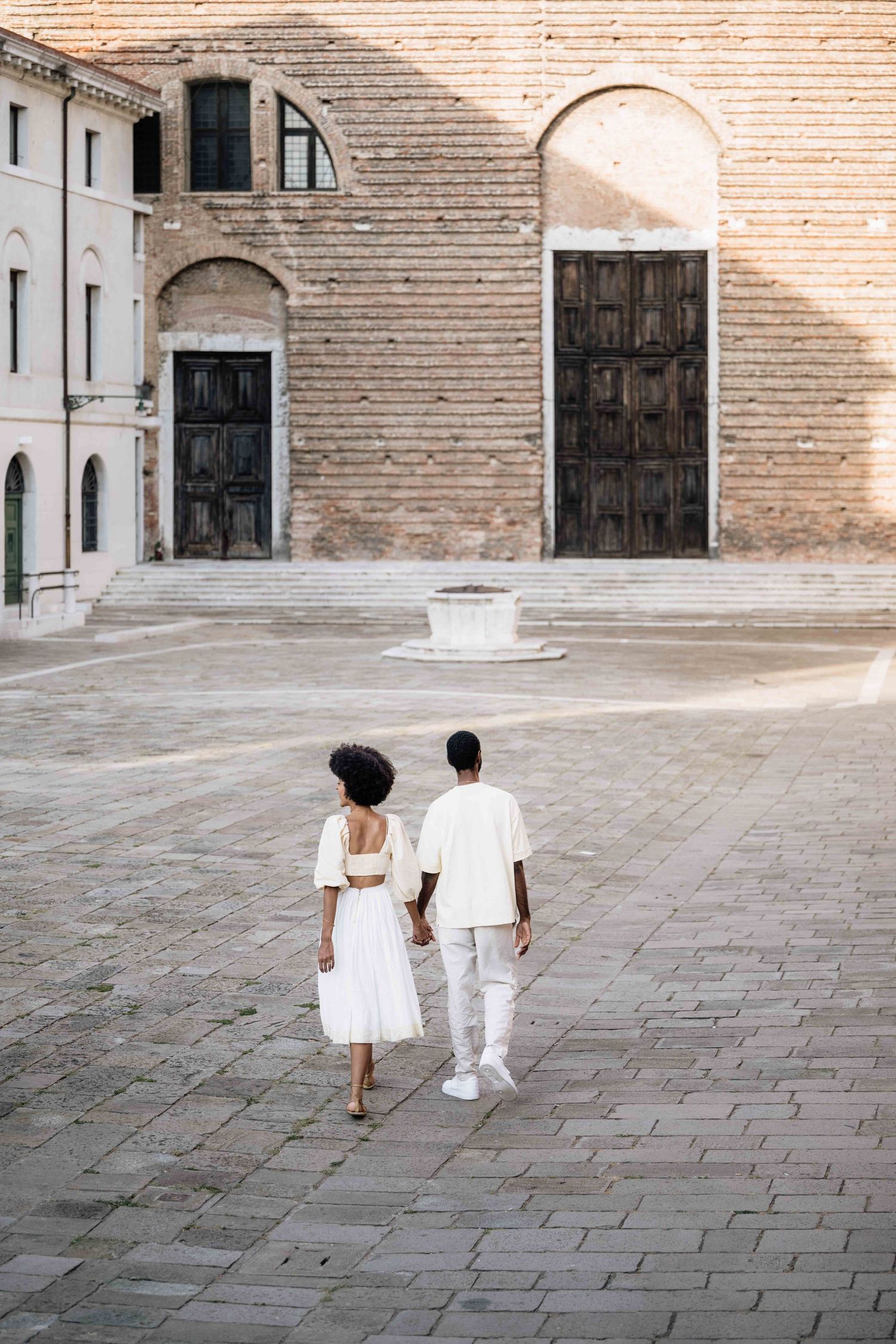 Two people in white outfits walking across a large stone courtyard with historic architecture.