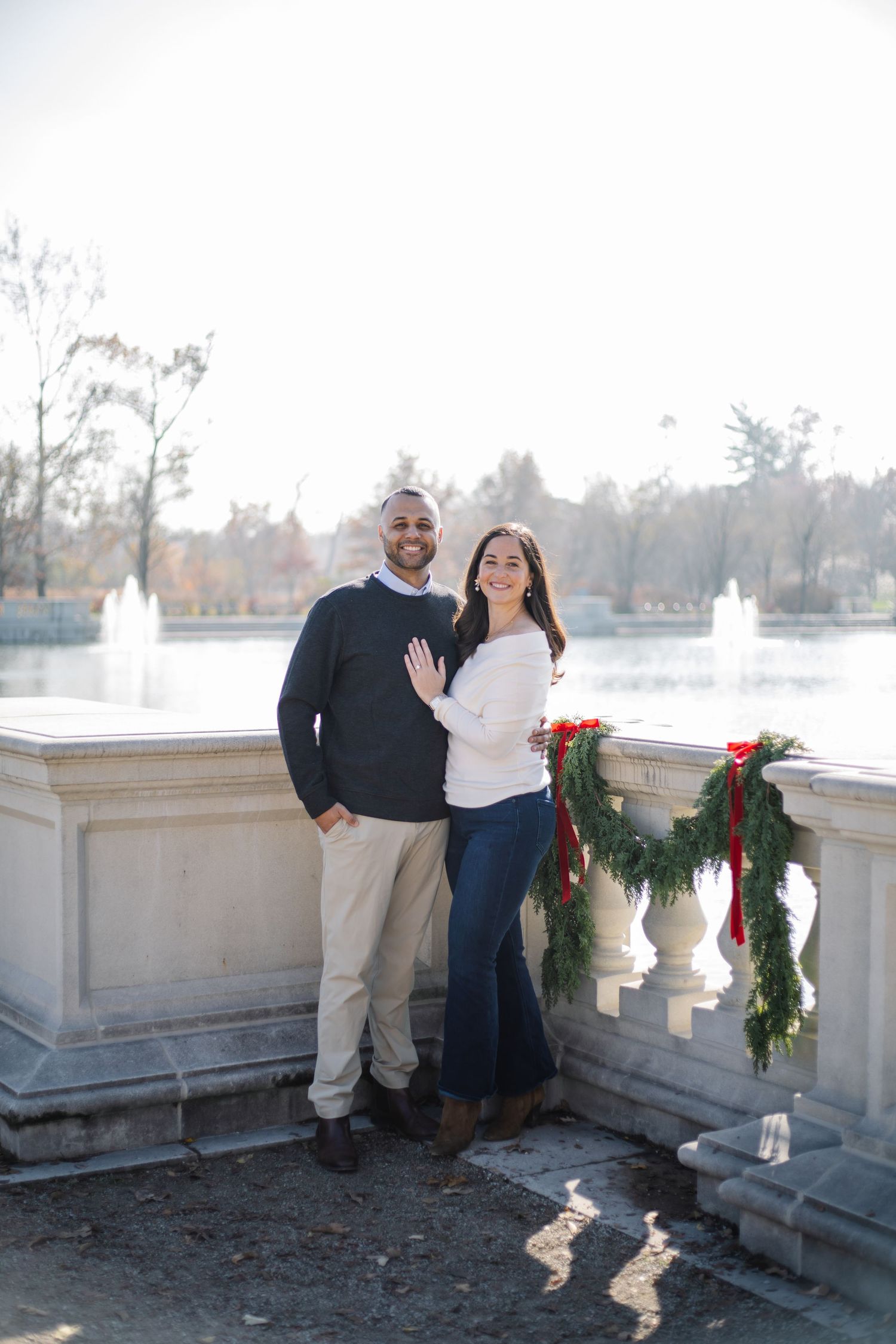 Happy couple poses together on a balcony with winter decorations and evergreen garland with red bows.