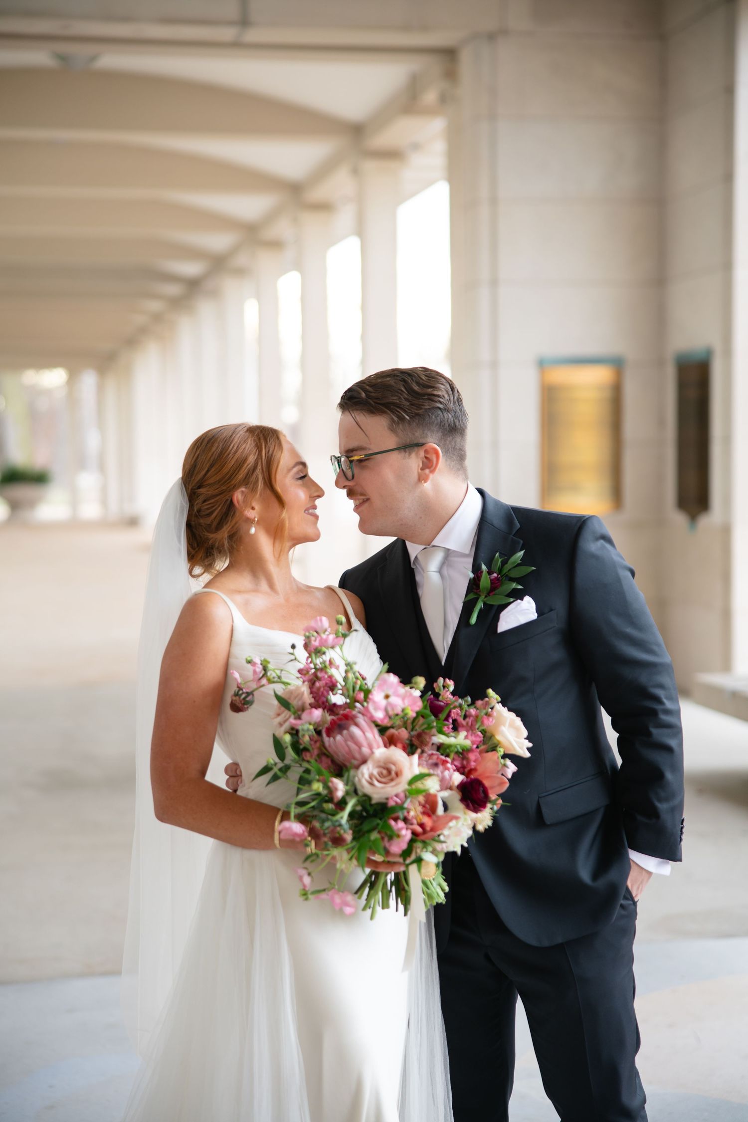 A romantic wedding couple shares an intimate moment in a bright hallway with white columns, holding a pink floral bouquet.