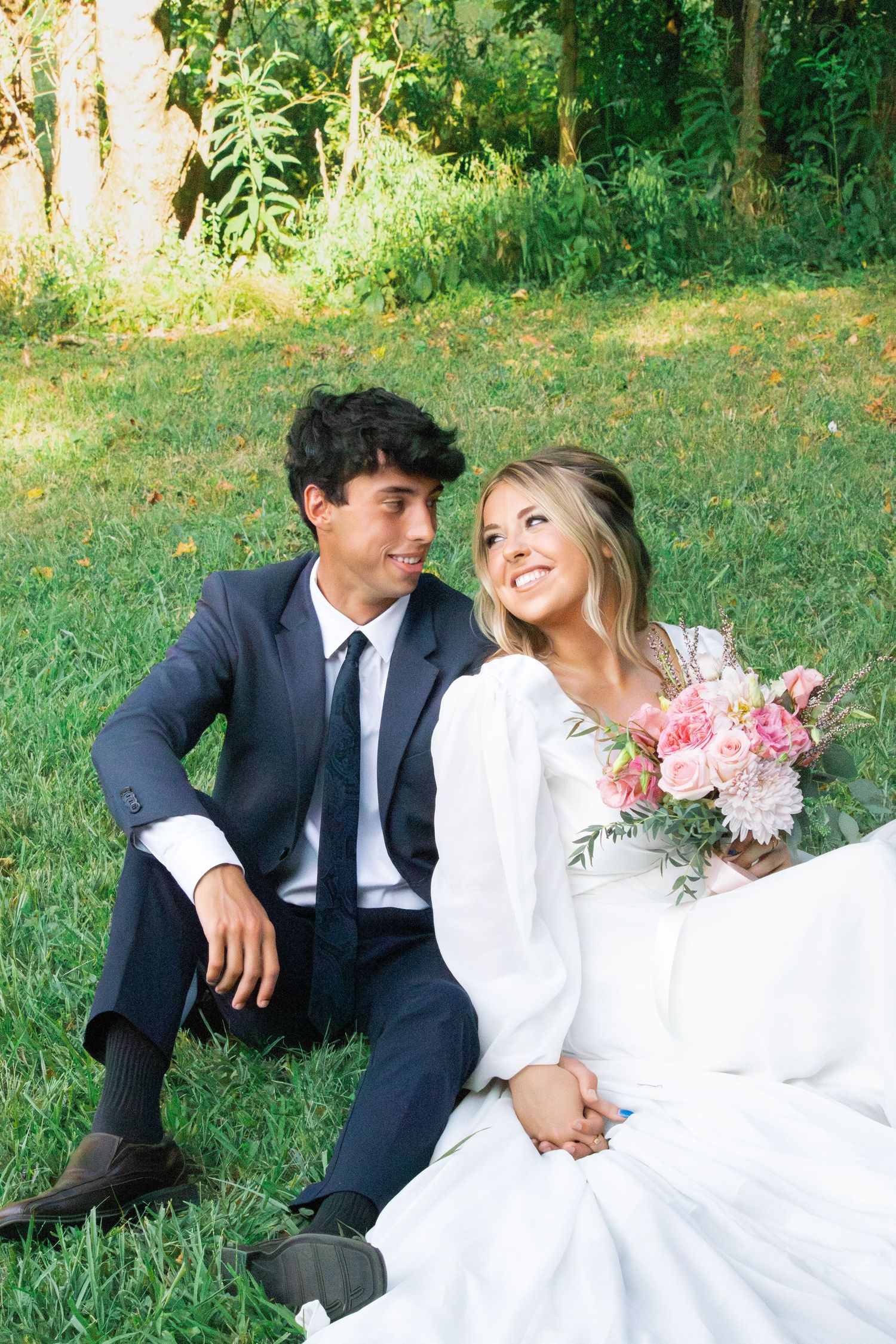 A bride and groom share intimate moments while sitting on grass in a garden setting.