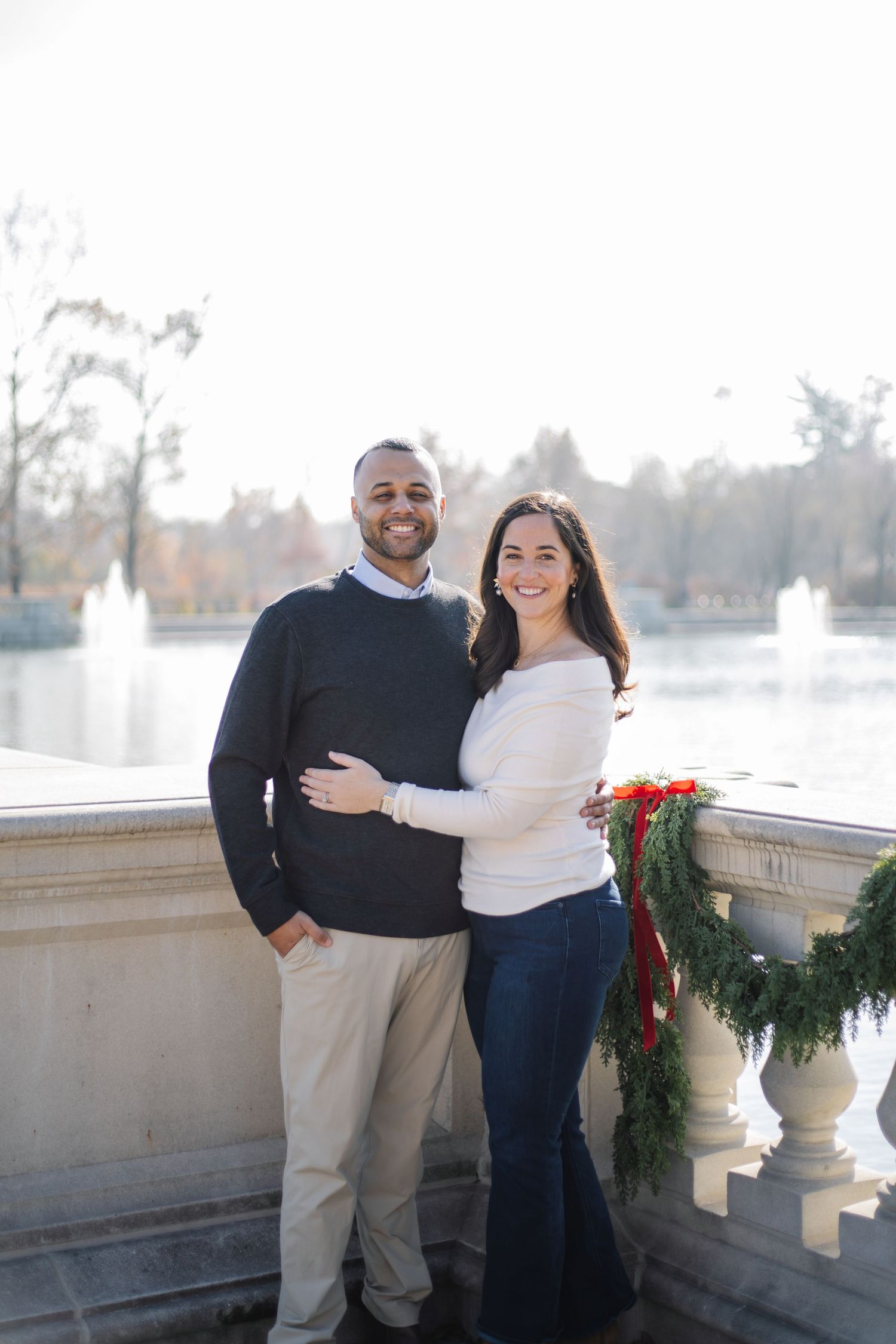 A couple poses together on a balcony with decorative stone railings and holiday decor.