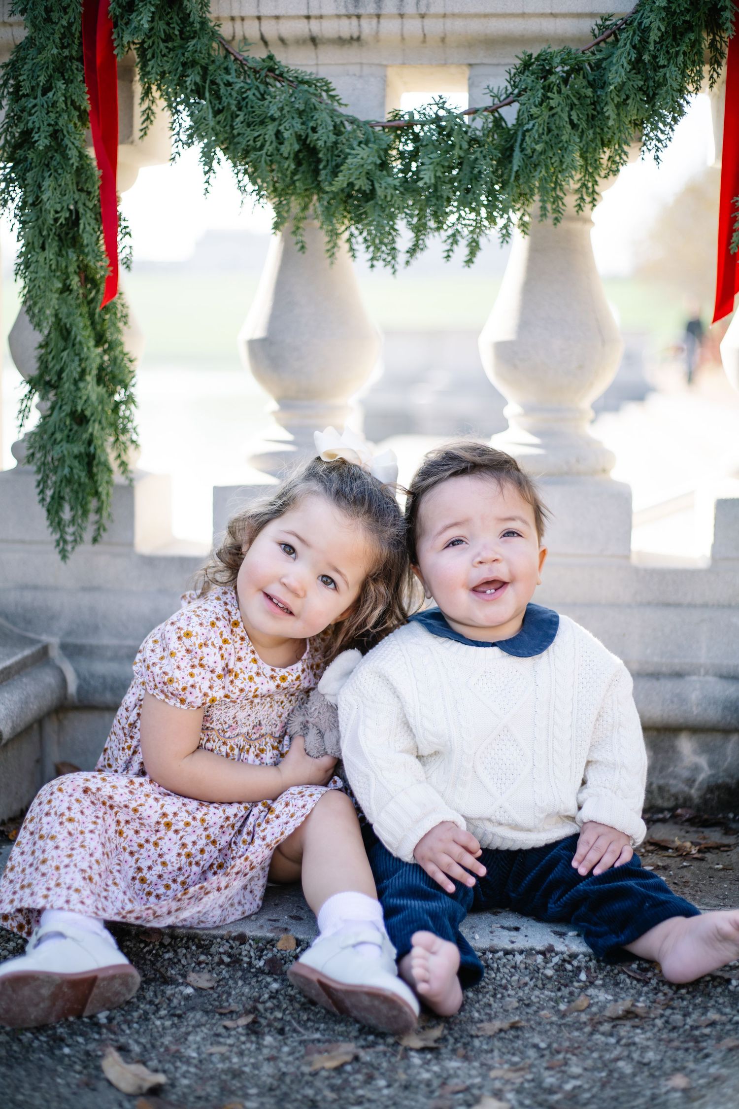 Two young children in casual attire sit together and embrace while smiling outdoors.