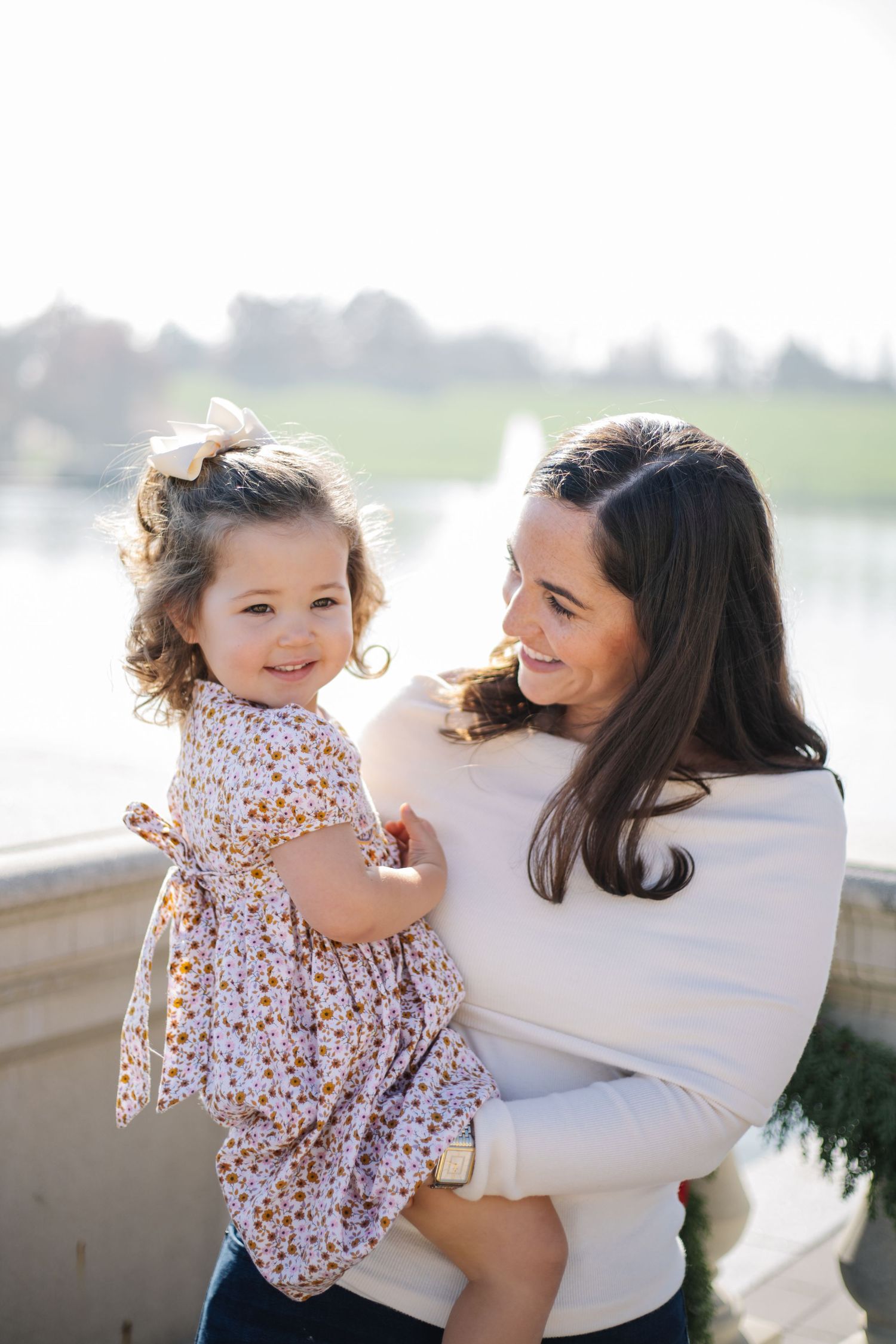 A sweet family moment captured outdoors with a small child in a floral dress being held against a bright background.