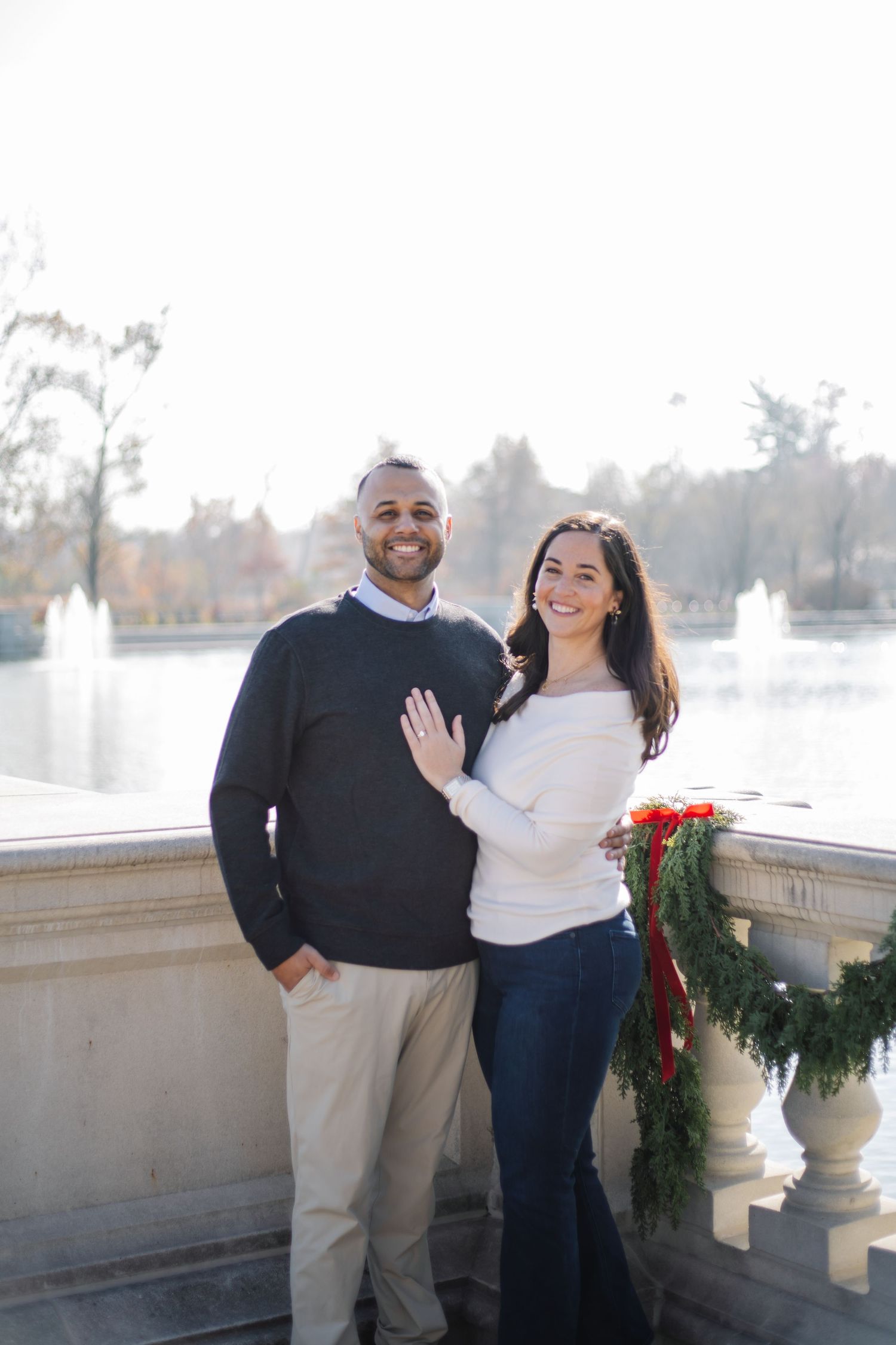 A couple poses together on a stone balcony decorated with holiday greenery during a winter engagement photoshoot.
