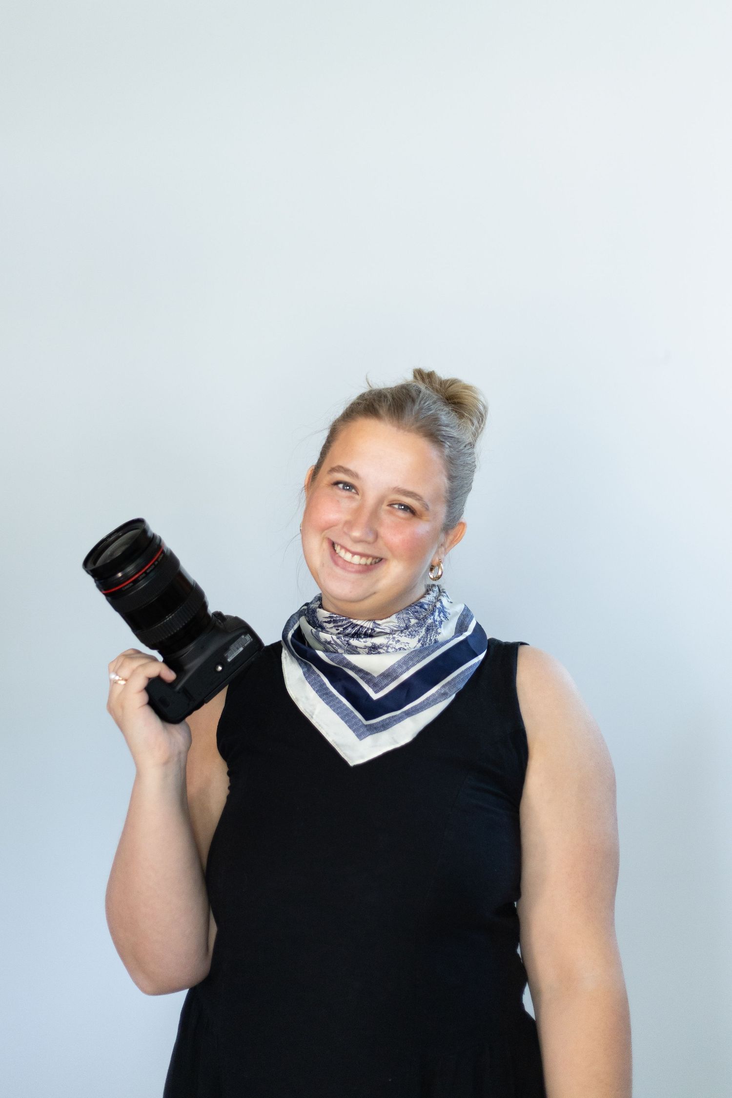Professional photographer in navy vest and bandana holding camera equipment against white background.