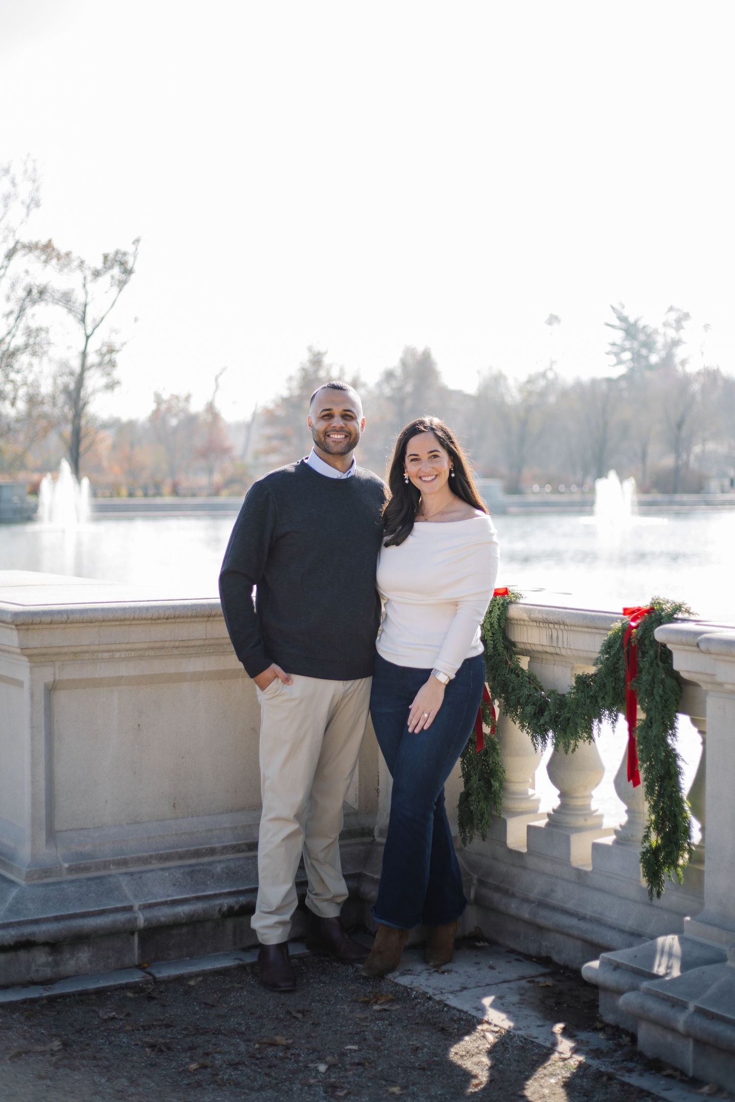 A couple poses together on a winter day near a stone balustrade decorated with holiday wreaths.