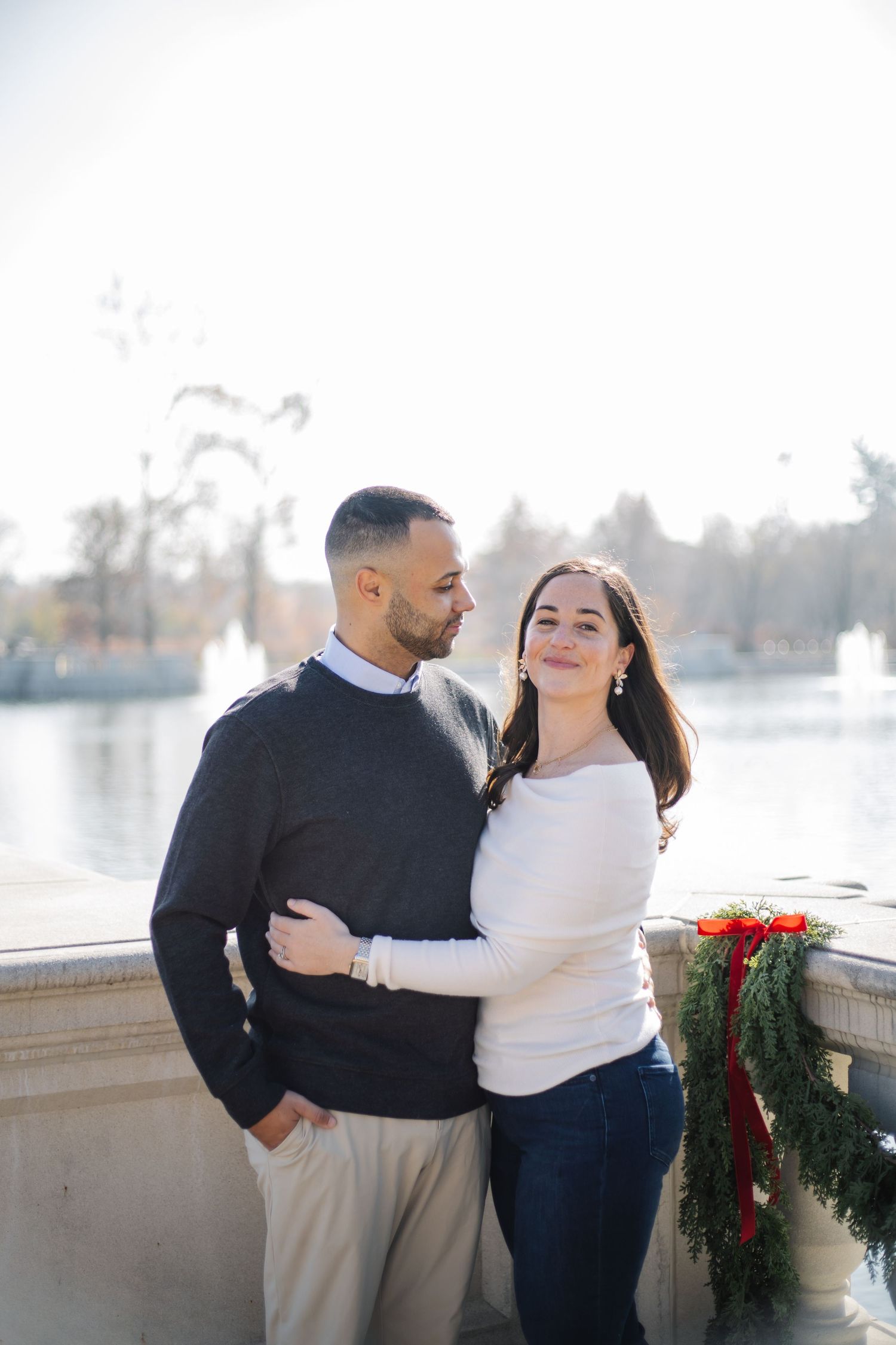 Stylish couple poses together by stone railing with holiday garland and lake view.