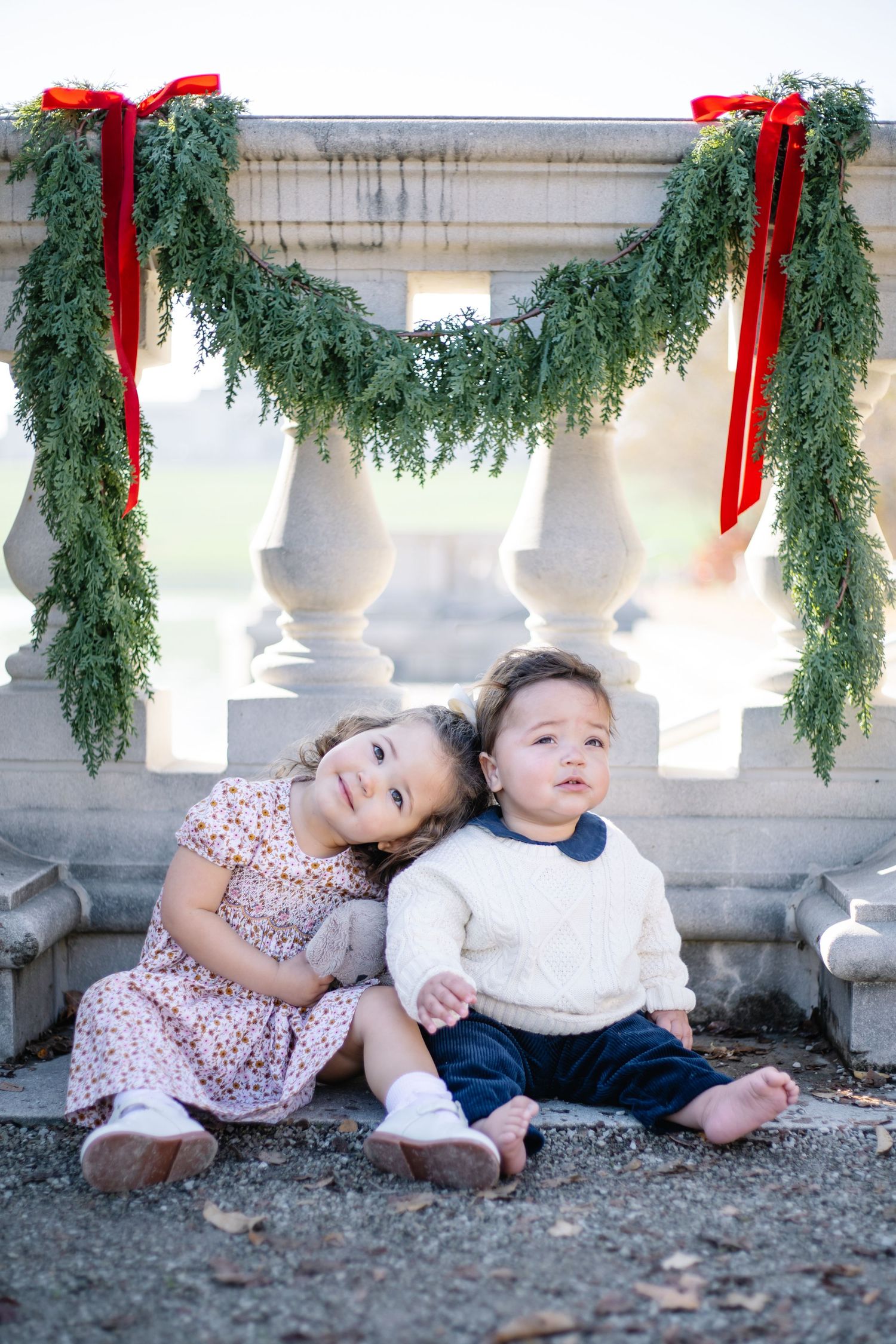Two children sit together under festive holiday garland with red bows on white stone columns.