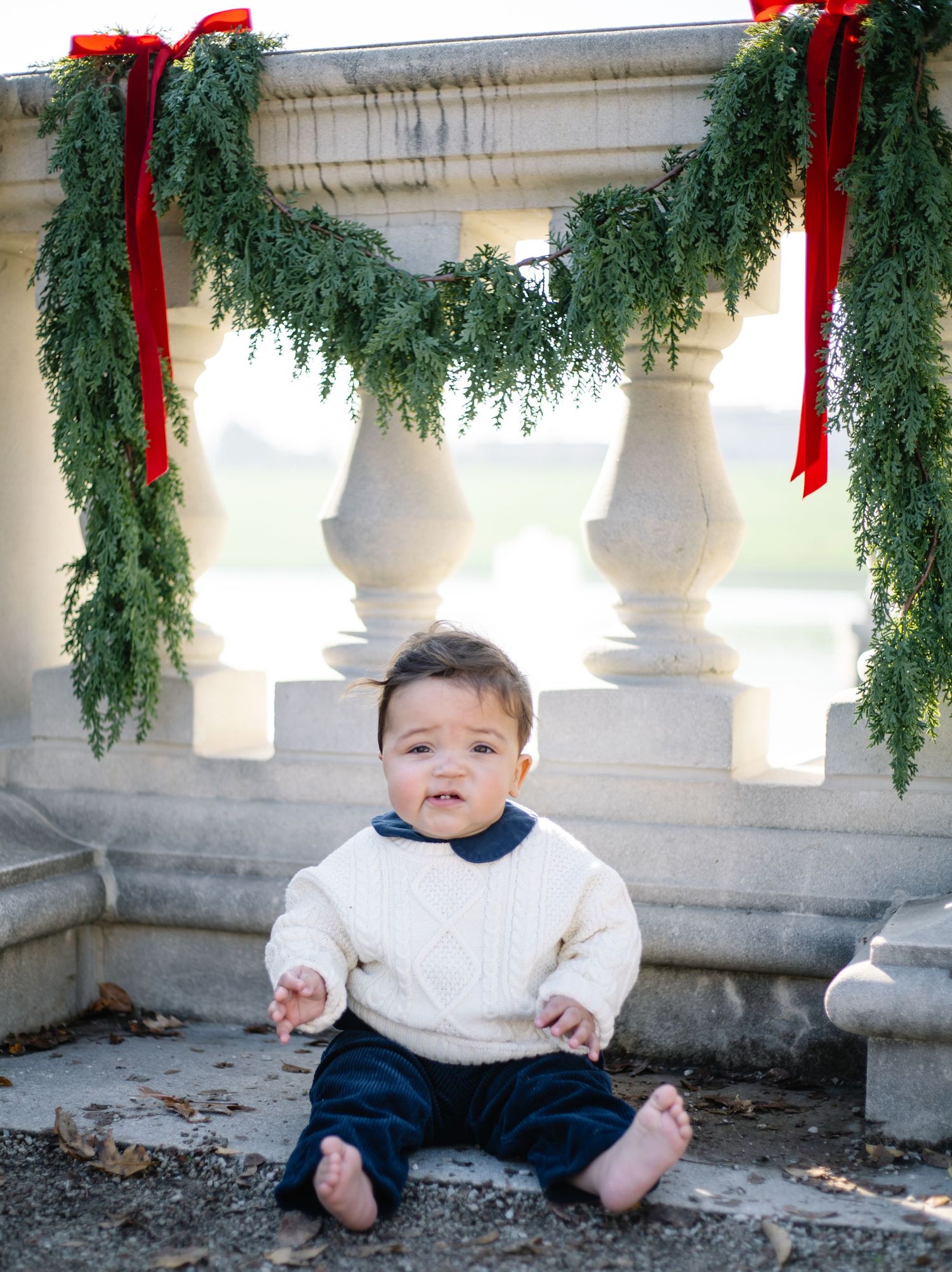 Baby in white sweater and navy pants sitting by stone railing decorated with holiday greenery.